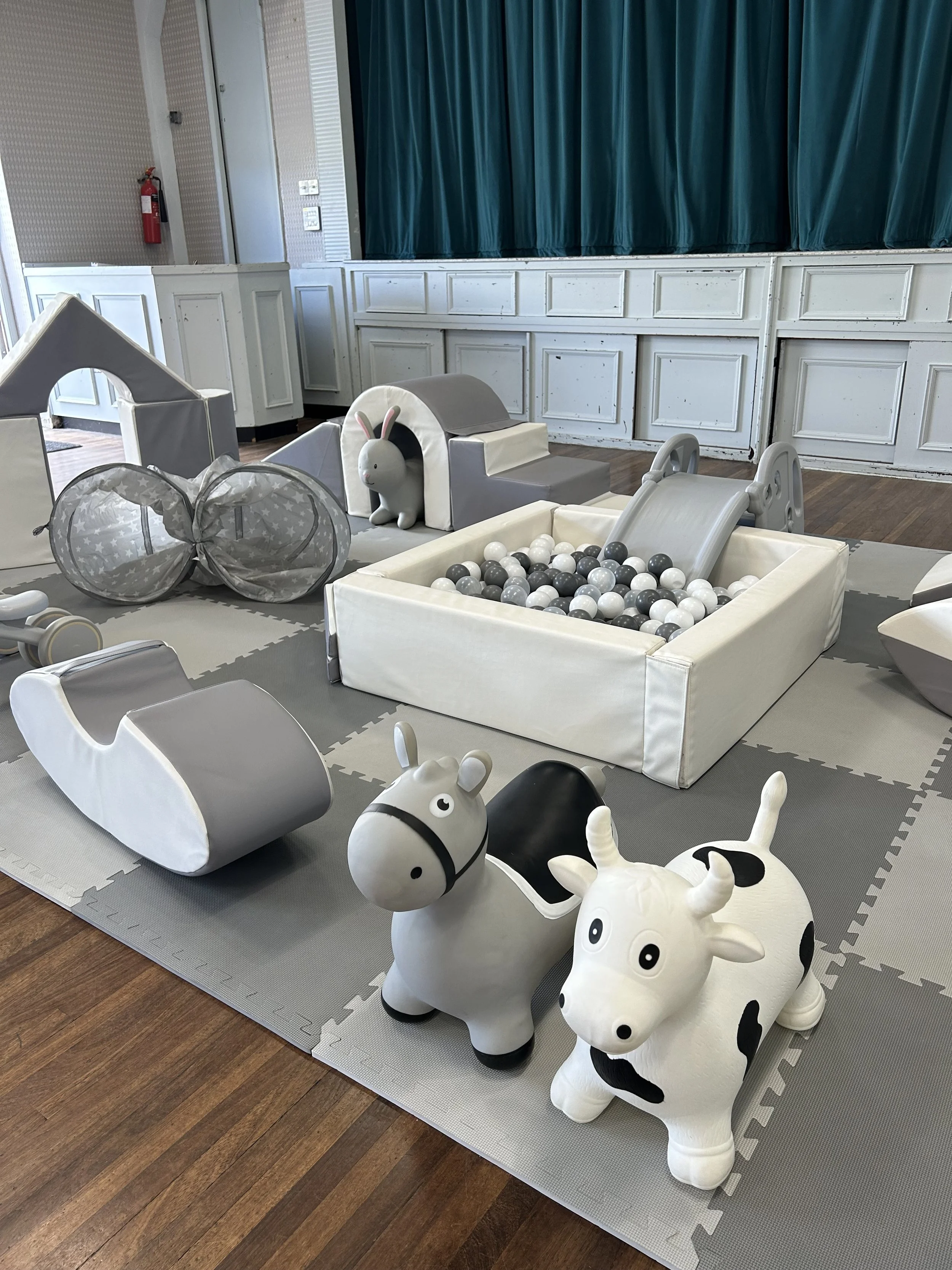 Children's play area with a ball pit, slide, stuffed animal toys, and soft play equipment, all in neutral tones of gray, white, and black.