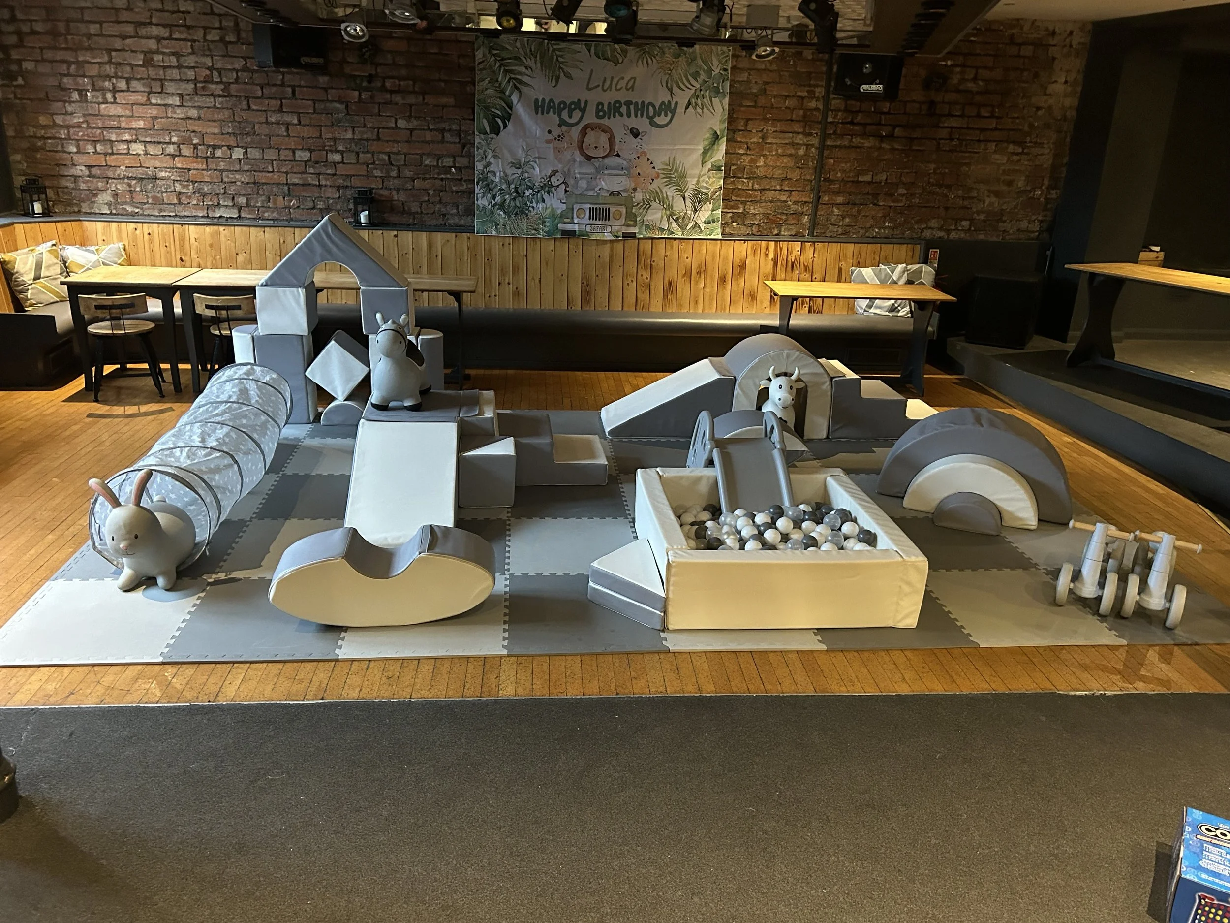 Children's indoor play area featuring soft play equipment such as a slide and animal-shaped structures, with a birthday banner on the brick wall reading 'Luca Happy Birthday' and decorated with jungle theme elements.