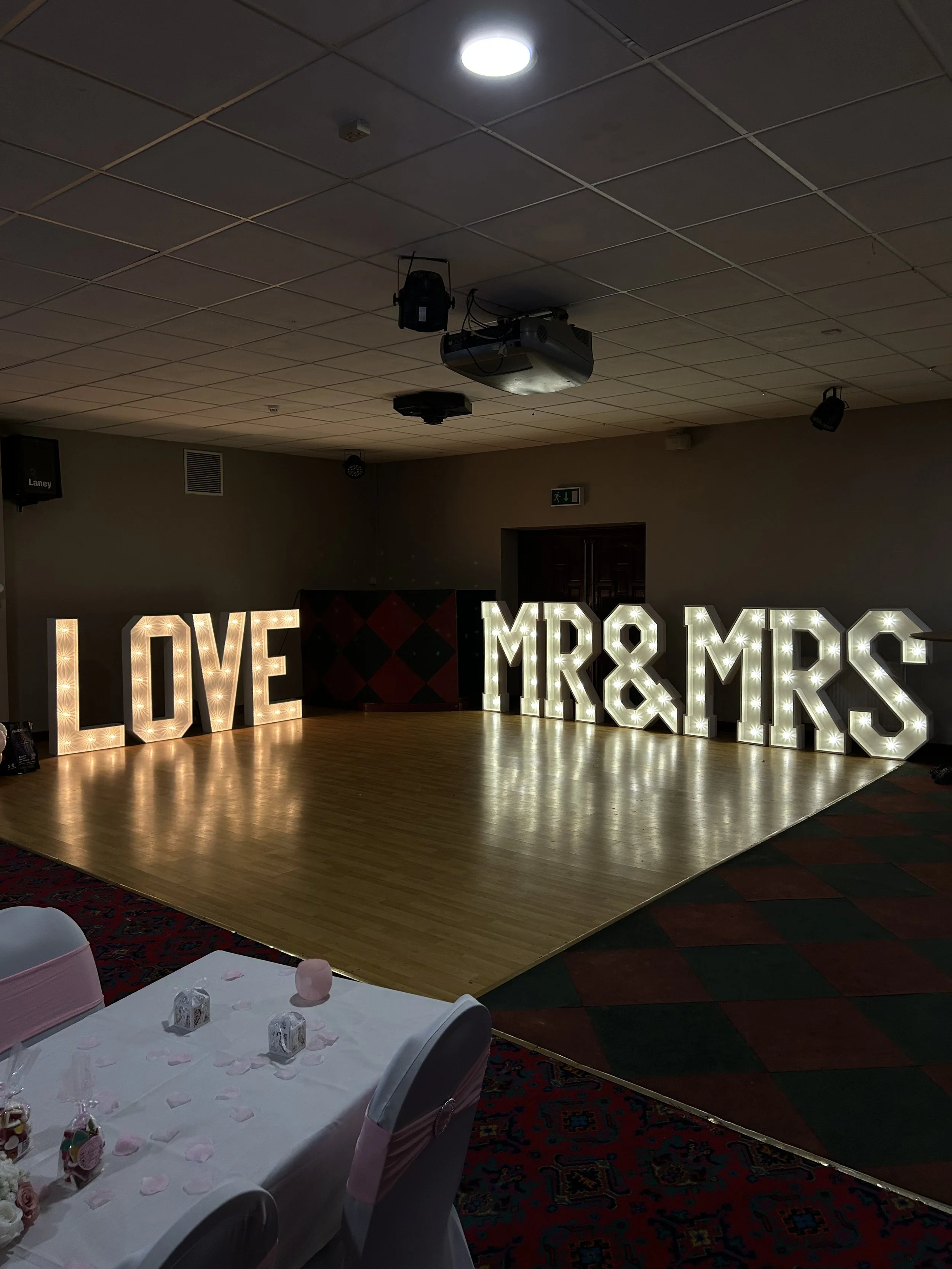 Decorated event space with illuminated large letters spelling "LOVE MRS & MR S" on a wooden floor, with tables featuring pink accents in the foreground.