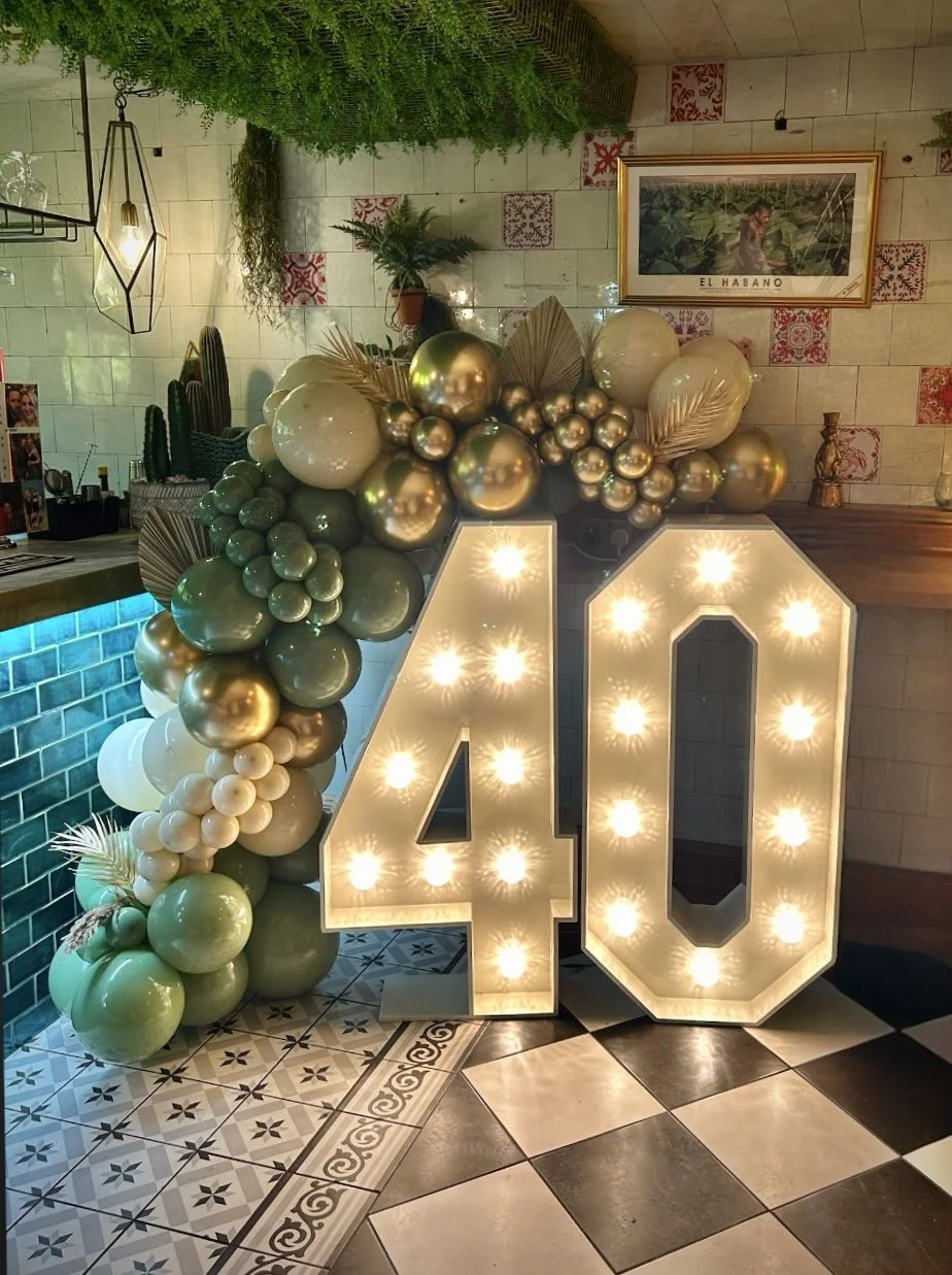 Decorative setup with large illuminated number 40, surrounded by gold, white, and pale green balloons. The background features a tiled wall, framed picture titled 'El Habano,' potted plants, and a hanging light fixture.