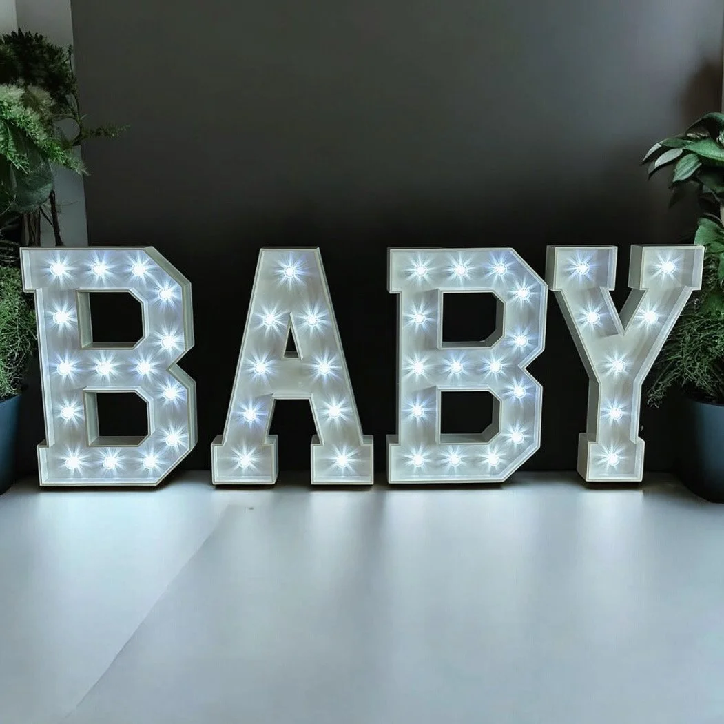 Decorative marquee spelling “BABY” with illuminated bulbs, placed on a white surface with green plants on both sides.