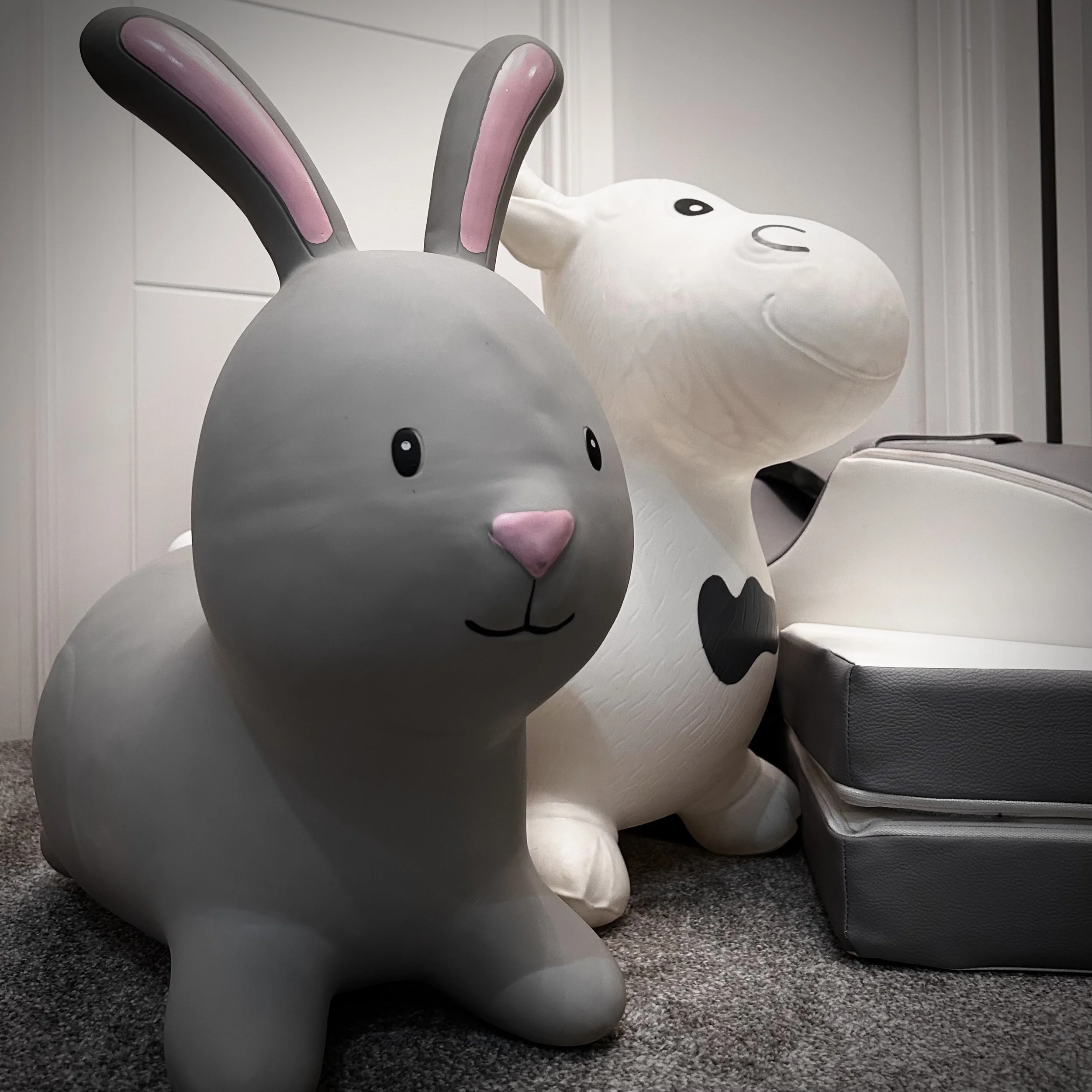 Two large bunny-shaped sculptures, one gray with pink ears and nose and the other white with a black heart on its chest, are placed on a carpeted floor next to a white wall and a grey cushioned seating area.