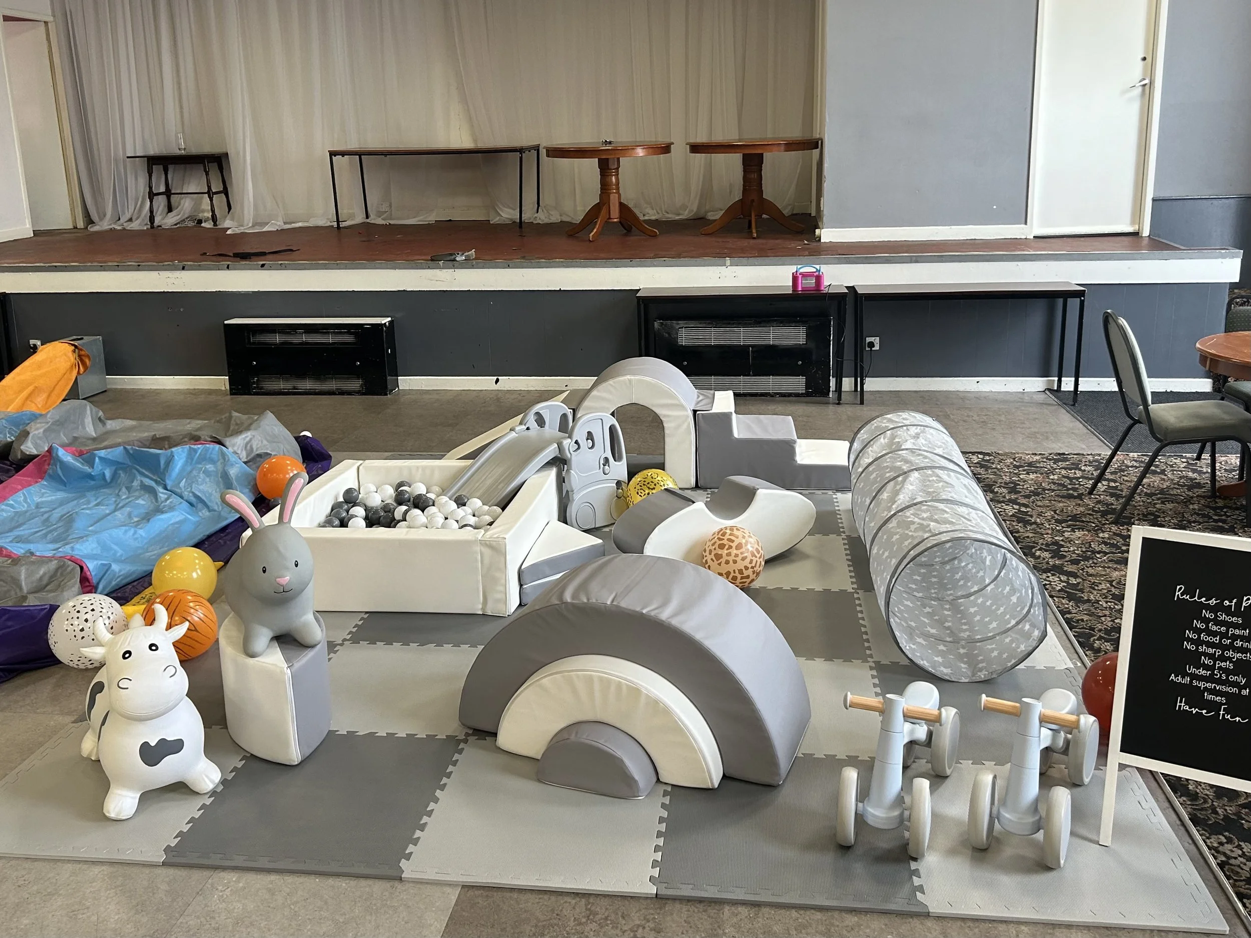 Children's indoor play area with soft play structures, toys, and a small stage in the background.