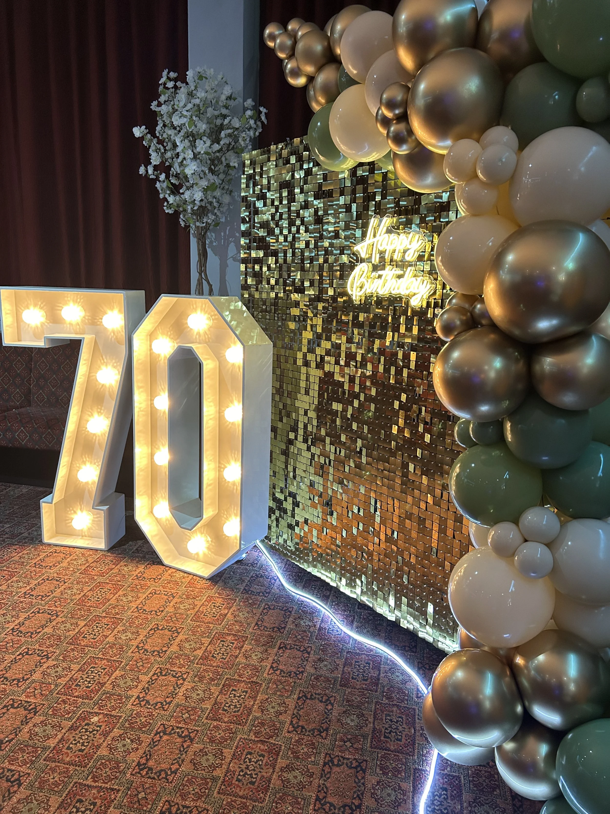 Decorative birthday setup with illuminated number 70, a balloon arch in gold, white, and green, a backdrop with small reflective tiles, a neon sign saying 'Happy Birthday,' a floral arrangement, and a multicolored carpet.