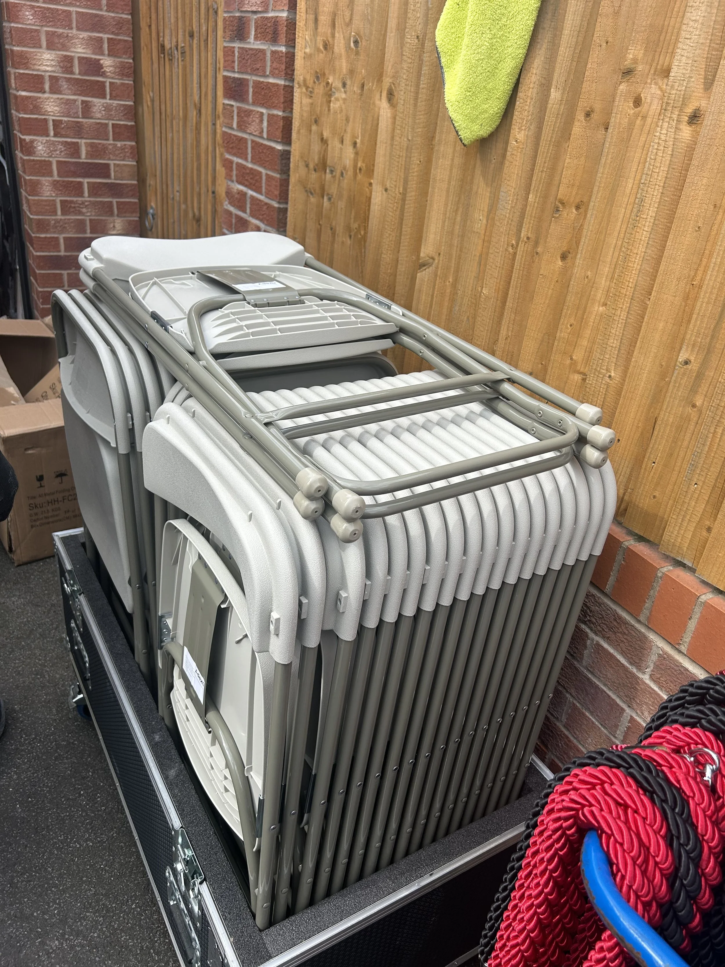 Stacked folding chairs stored outdoors against a brick and wooden fence, with a box and red and black ropes nearby.