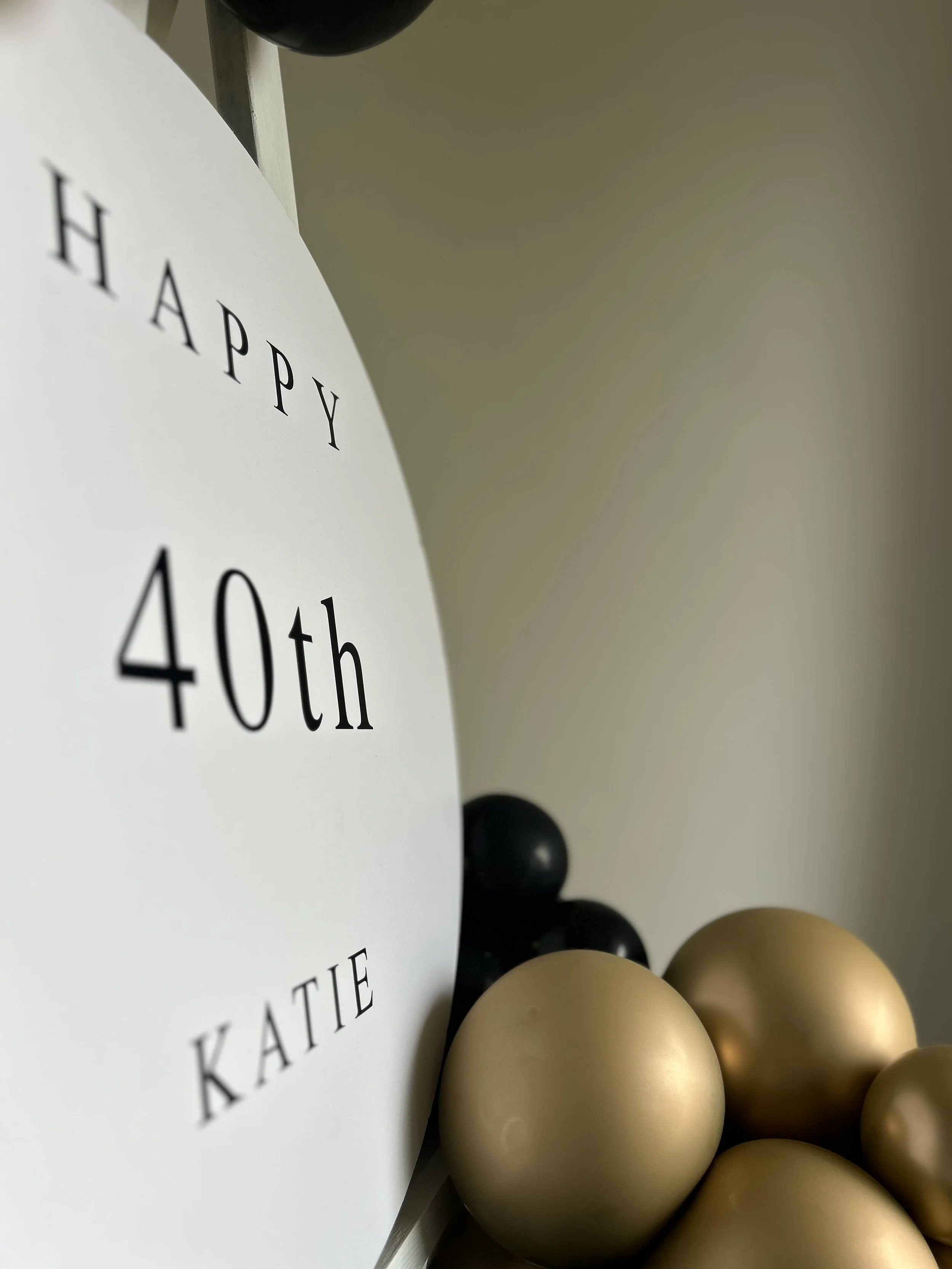Close-up of a white circular sign with black text reading 'Happy 40th Katie,' surrounded by black and gold balloons.