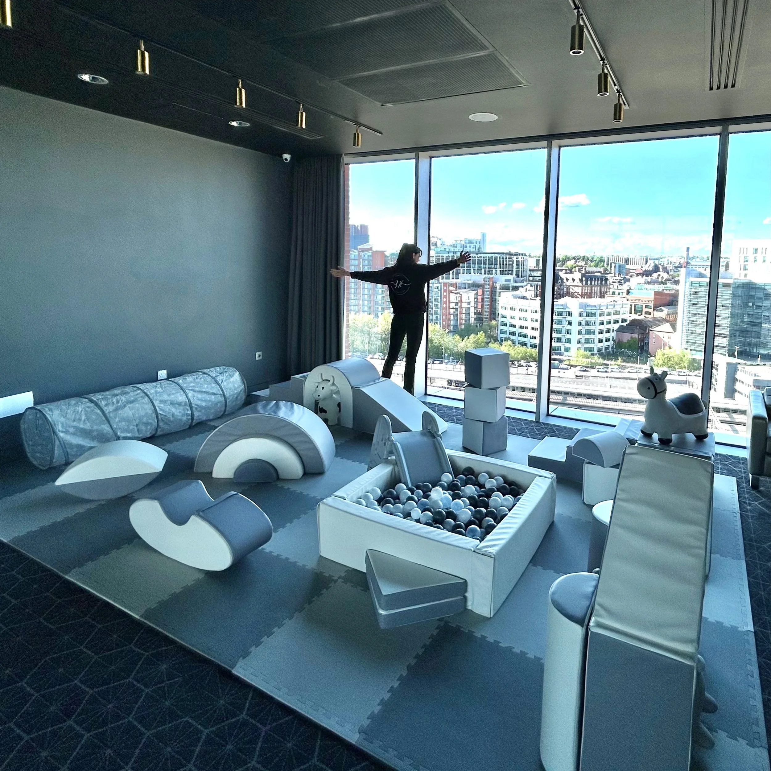 Indoor playroom with soft foam climbing structures, a ball pit, and a woman looking out the large window at a cityscape.