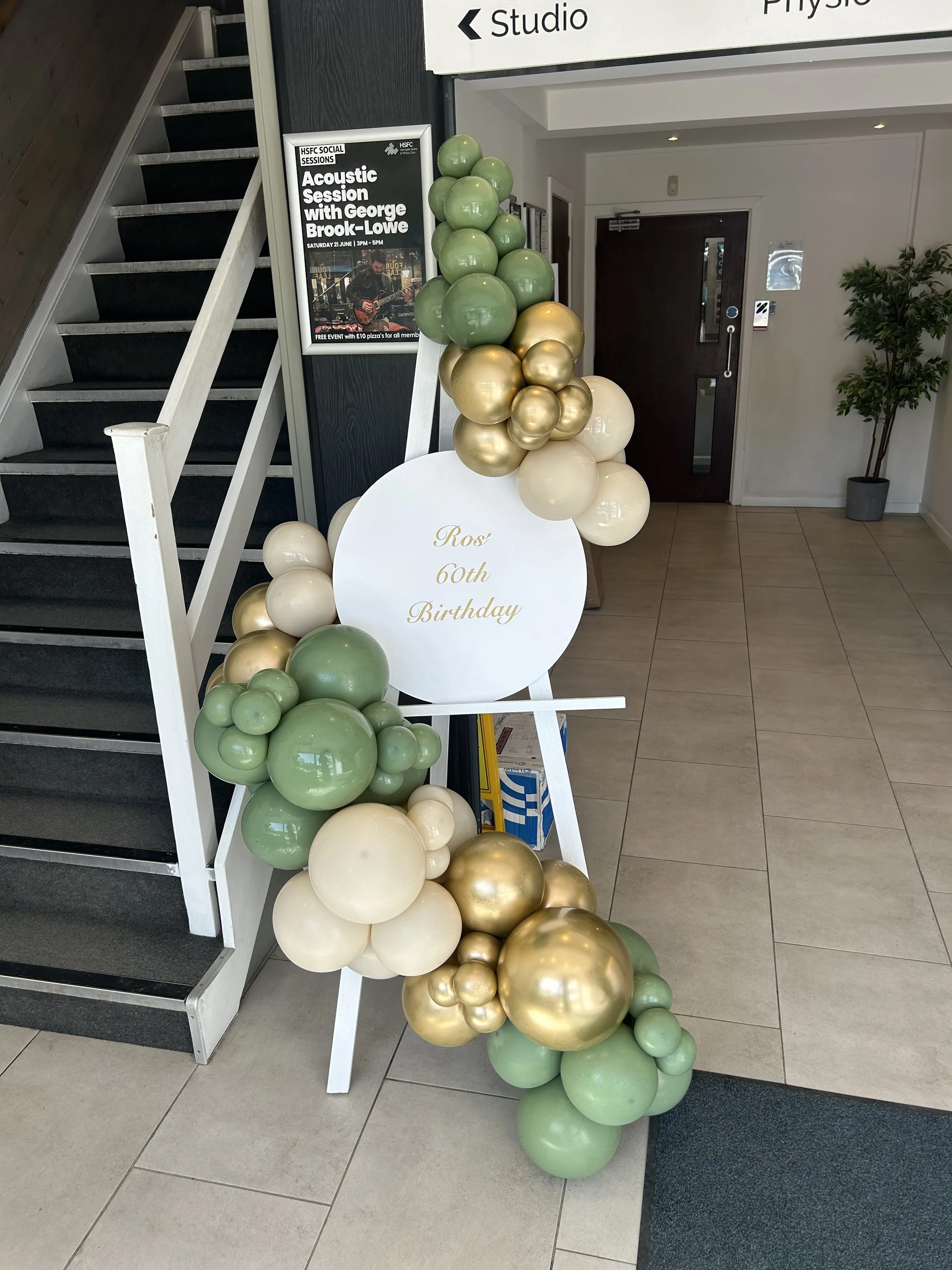 Balloon arrangement with green, cream, and gold balloons, with a white round sign that reads "Ros 60th Birthday".