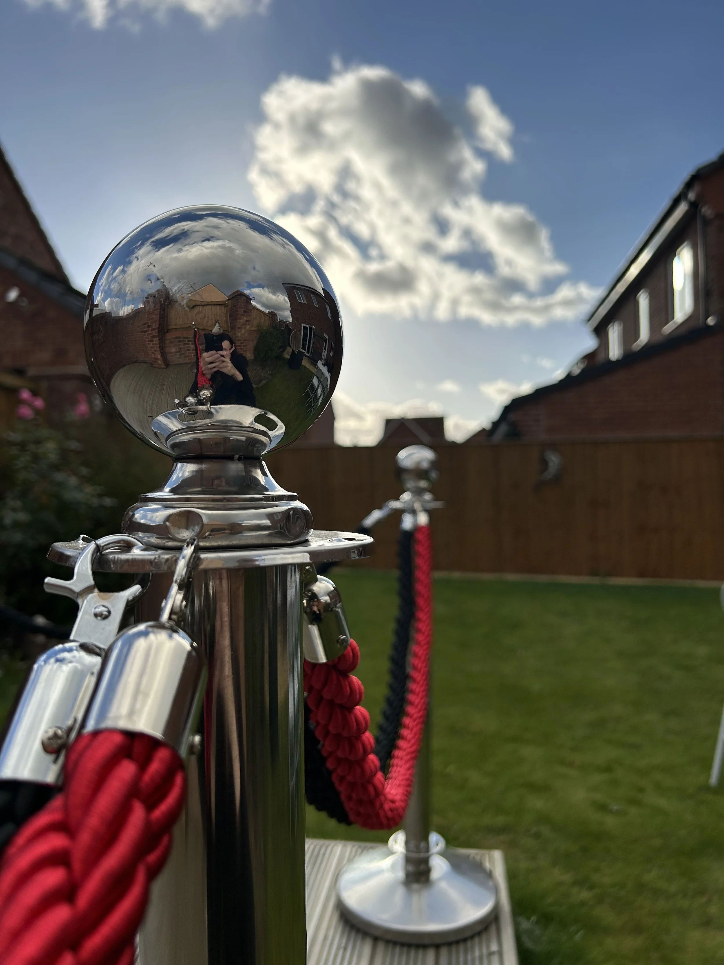 Close-up of a polished metal post with a reflective sphere on top, showing the person taking the photo, a clip, red and black ropes, and a backyard with a cloudy sky.