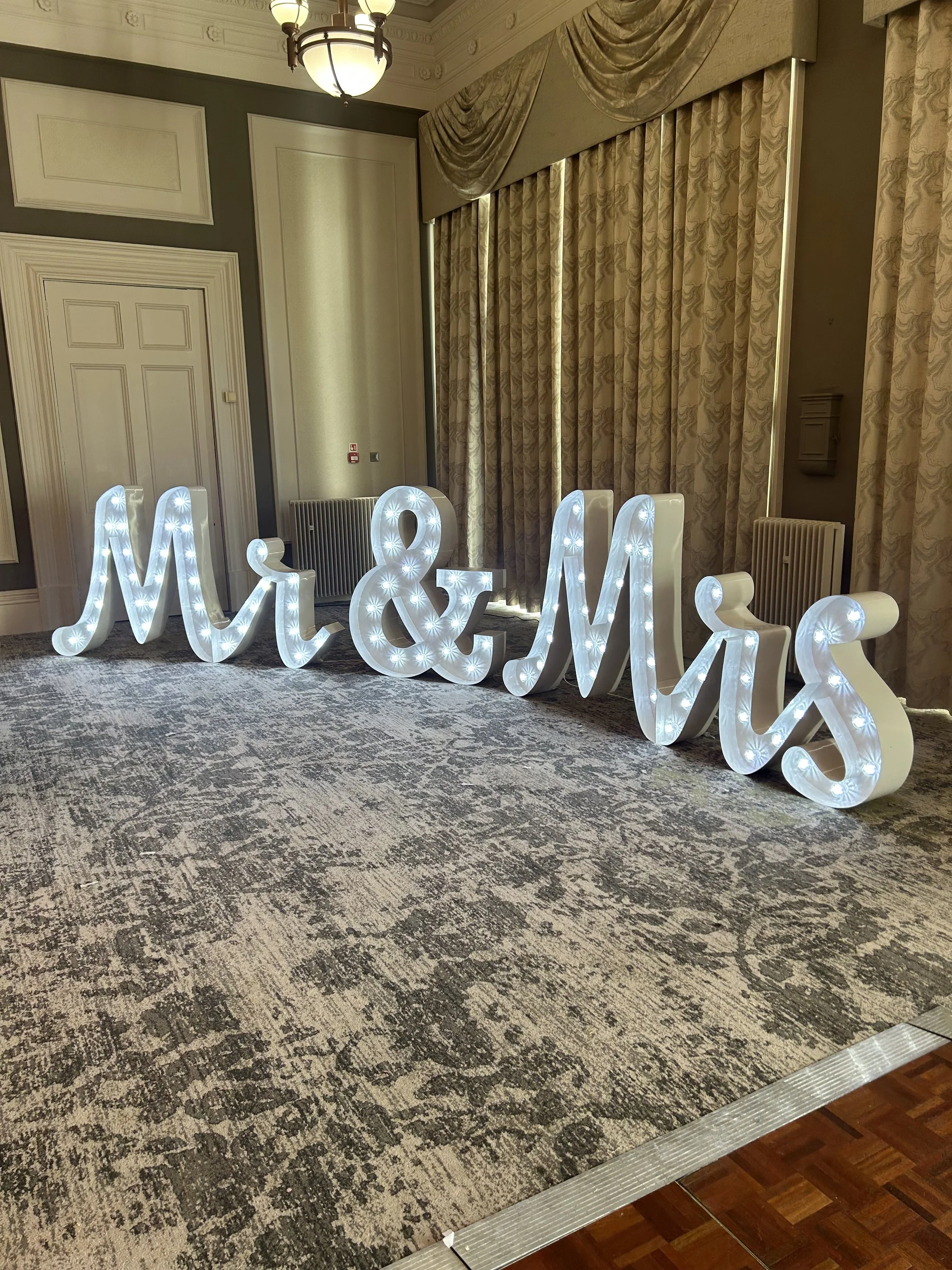 Large illuminated sign spelling 'Mr & Mrs' with white lights inside, placed on a patterned carpet in an elegant room with high ceilings, ornate curtains, and vintage decor.
