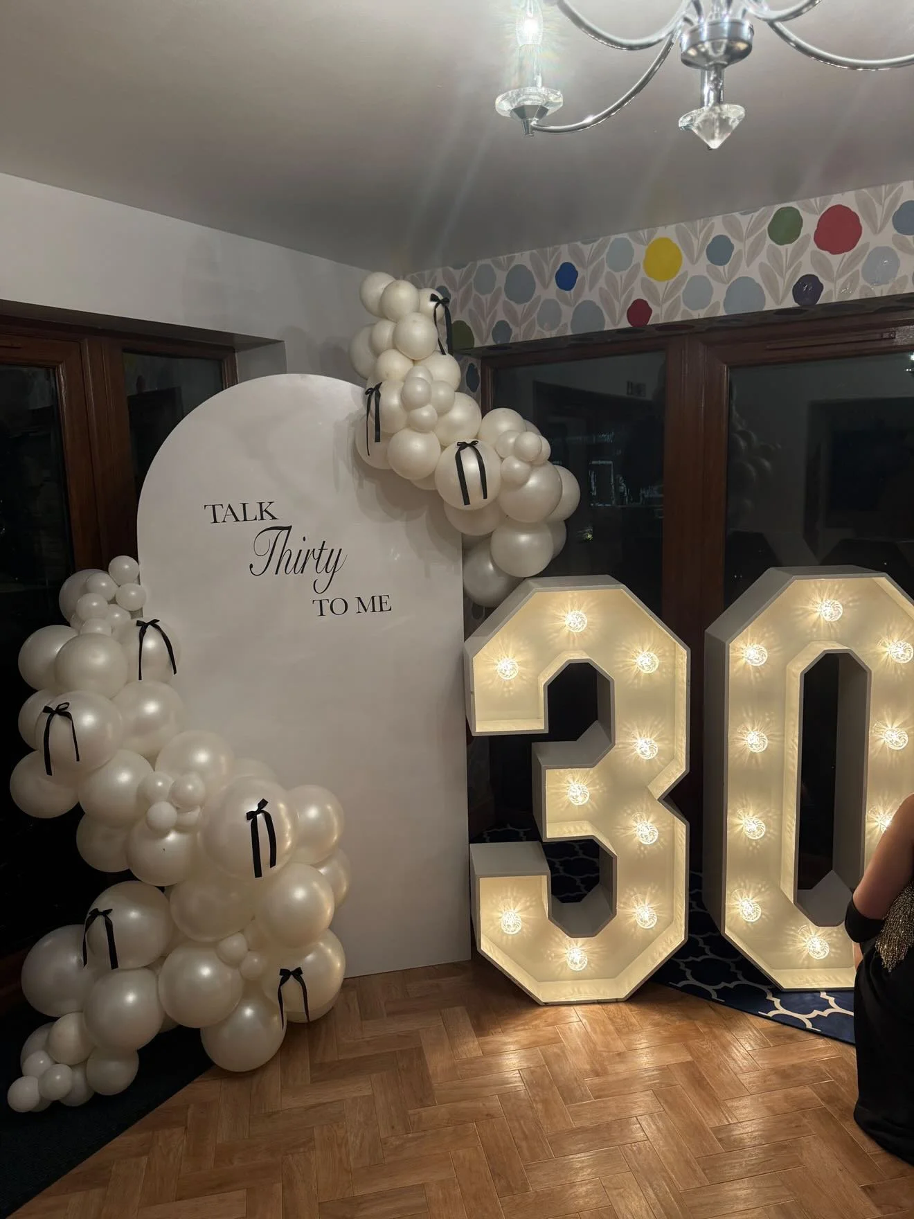 Decorations for a 30th birthday celebration featuring a large white backdrop with the phrase 'Talk Thirty to Me,' clusters of white balloons with black ribbon accents, and big number '30' illuminated with bulbs in a room with wooden floors and a colo