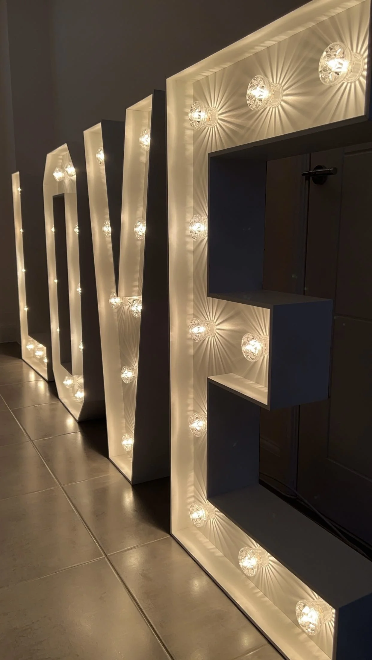 Large decorative sign spelling "LOVE" with illuminated bulbs around each letter, placed on a tiled floor against a dark wall.