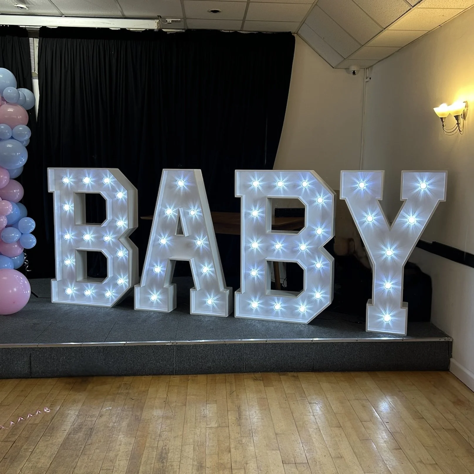 Large illuminated letters spelling 'BABY' on a small stage, with pink and blue balloons to the side, at an indoor celebration.