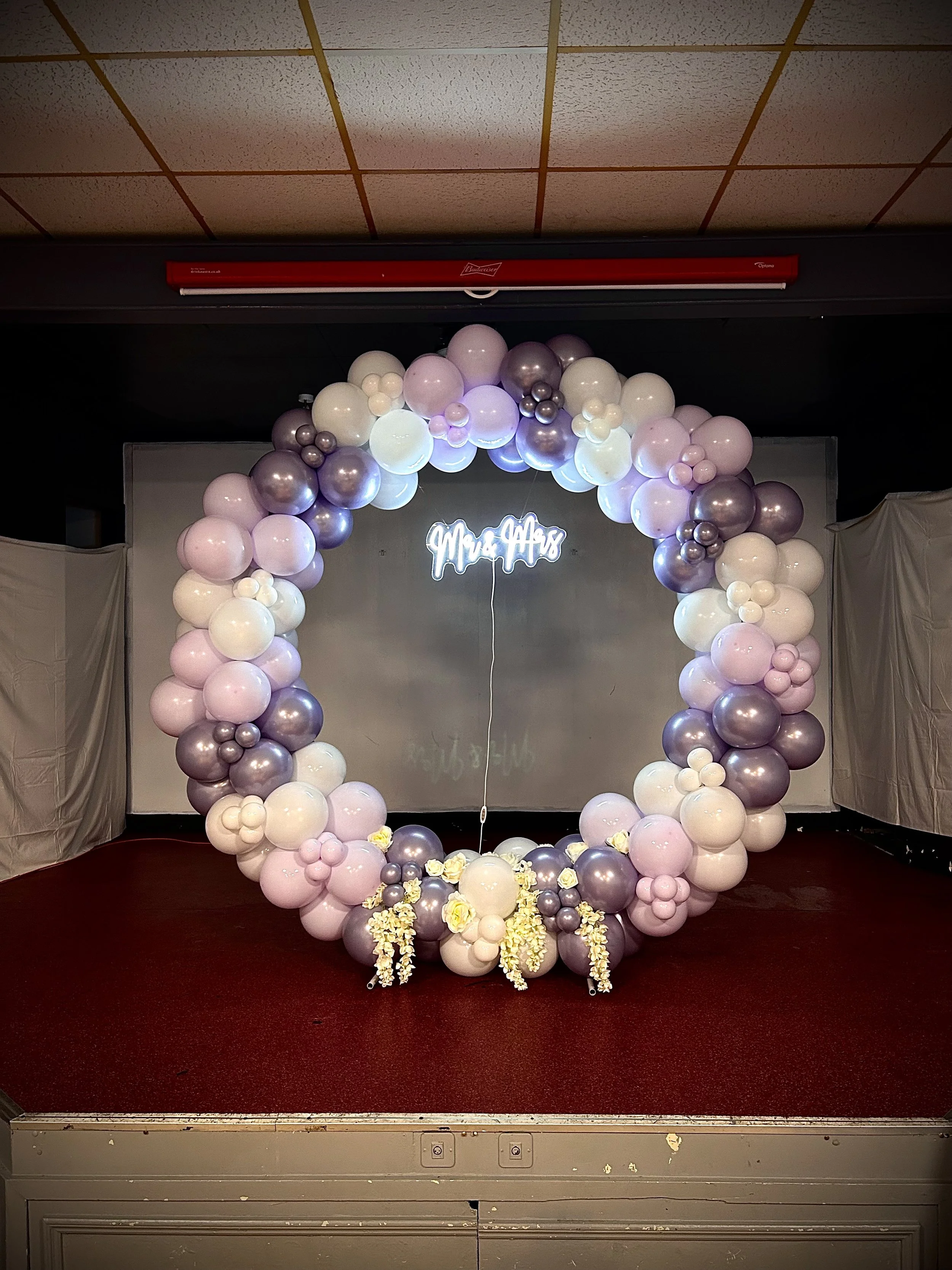 A circular balloon garland in shades of white, pink, and purple with some floral accents surrounds a neon sign that reads 'Mrs. Alice' in a decorated event space.