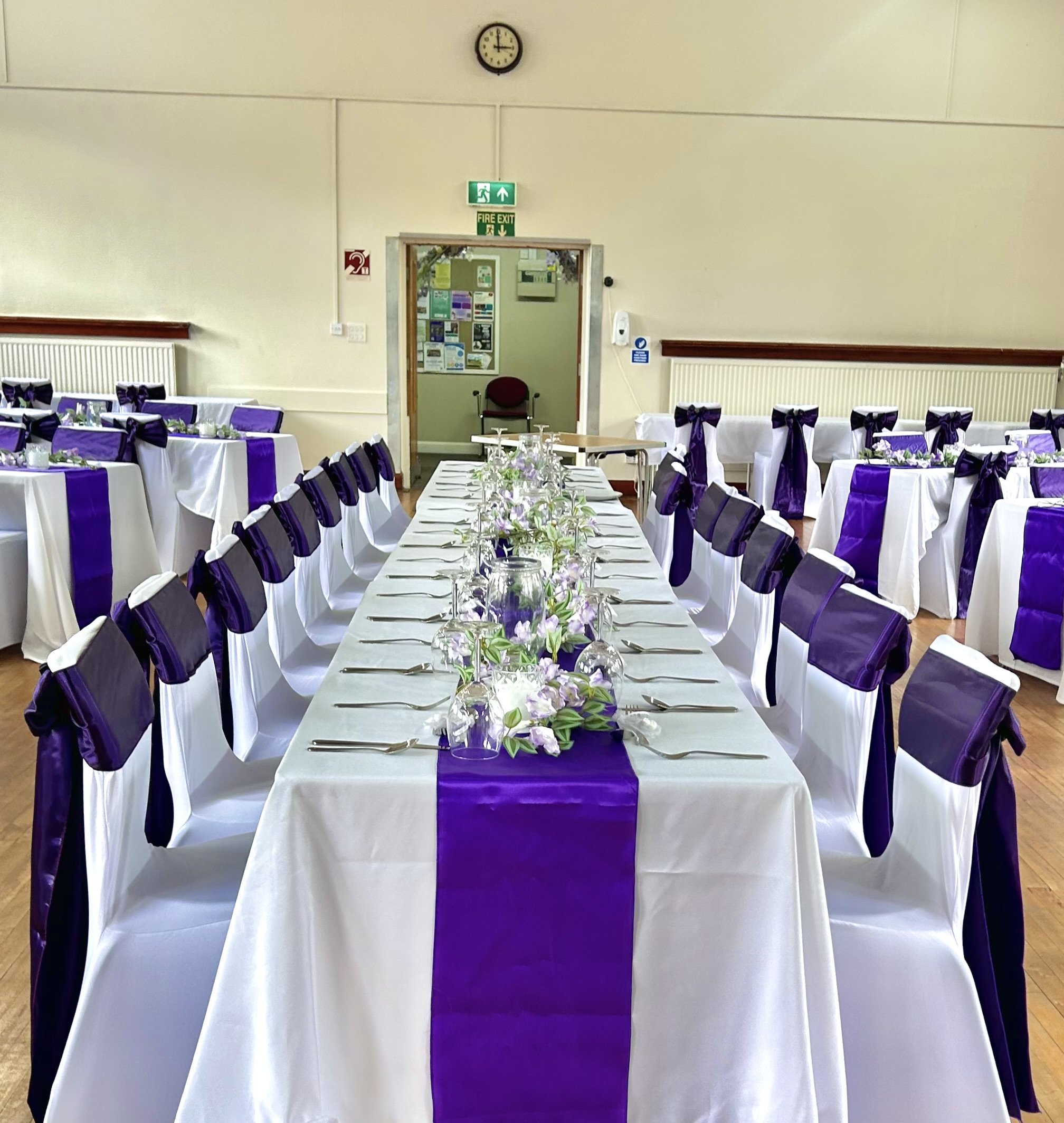 Event hall decorated with white and purple tablecloths, purple bows on chairs, and floral centerpieces. Fire exit and emergency equipment signs are visible on the wall.