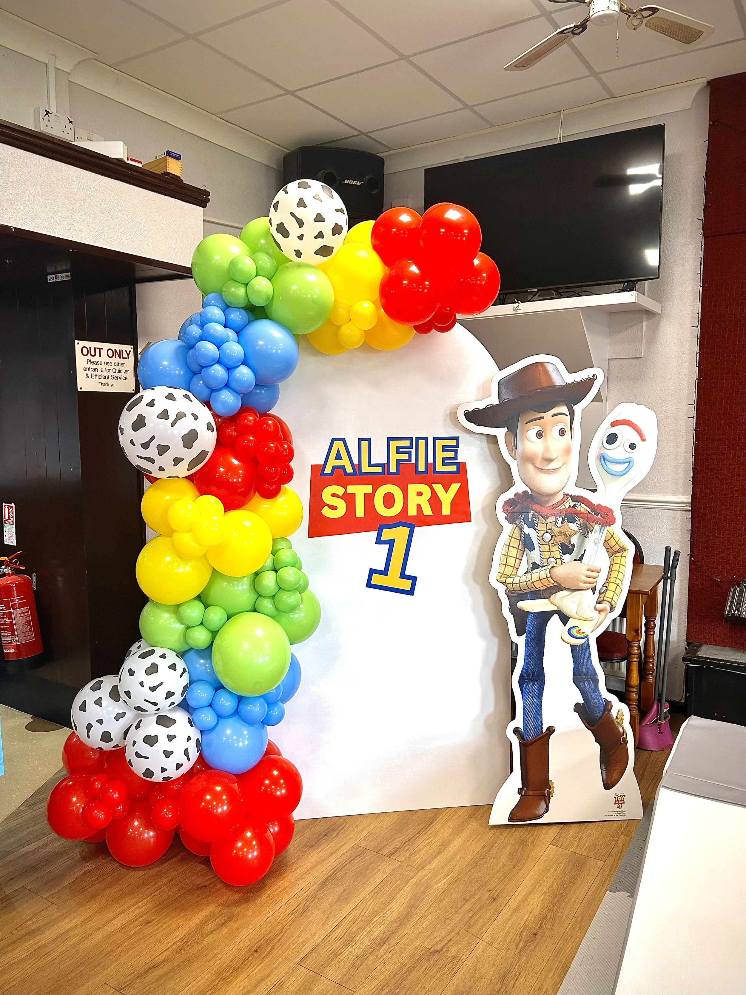 Decorative display for a child's first birthday party featuring a colorful balloon arch and a large cutout of Woody from Toy Story. The balloons are arranged in a gradient of red, yellow, green, blue, and contain some with cow print designs. The disp