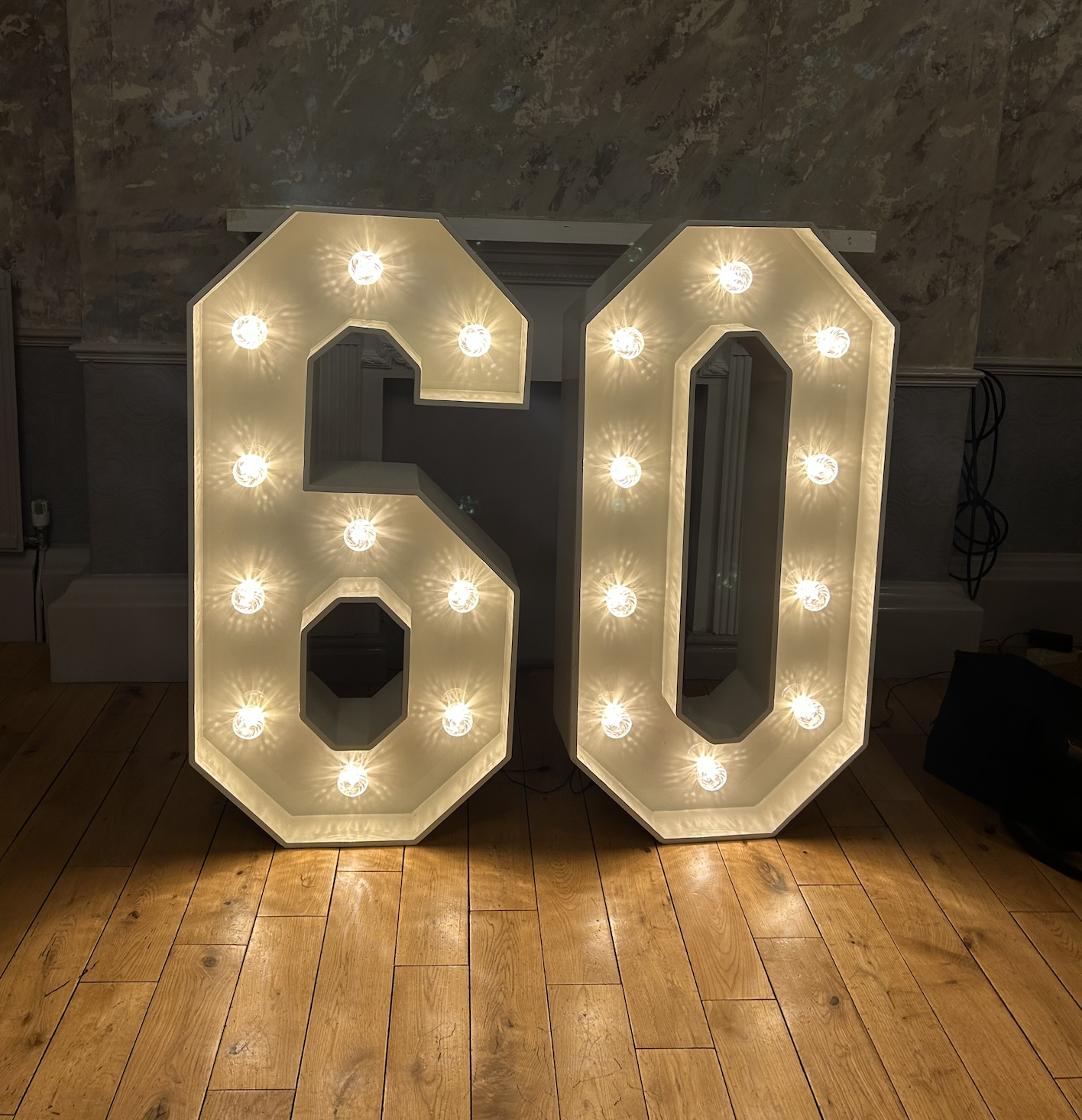 Illuminated marquee number 60 with bright bulb lights, standing on a wooden floor in front of a textured wall.