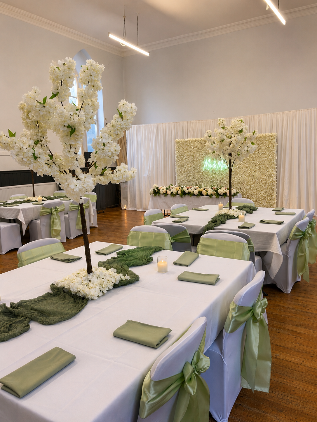 A decorated event space with white tablecloths, green napkins, and cream-colored chair covers tied with green ribbons. The tables have tall artificial trees with white flowers and green fabric runners. In the background, there is a floral backdrop wi