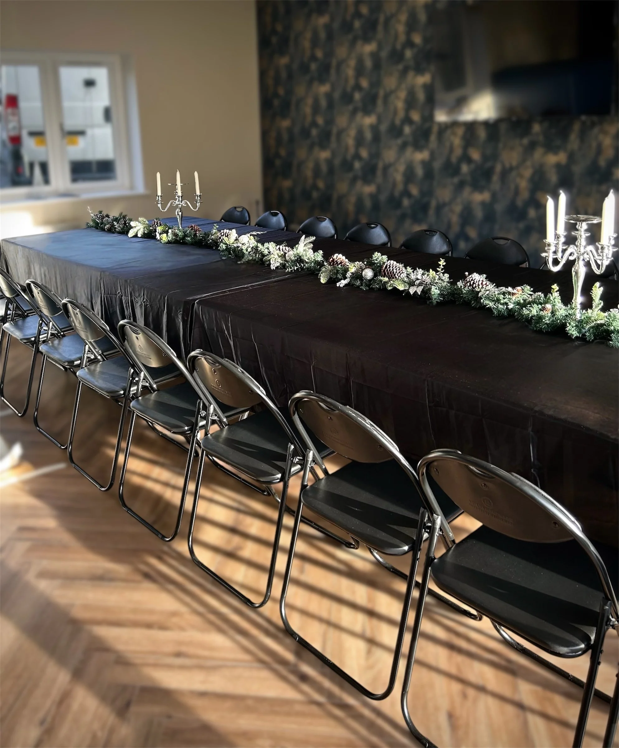A long table set for a formal event, covered with a black tablecloth, decorated with matching silver candelabras with lit white candles and holiday greenery with pinecones.