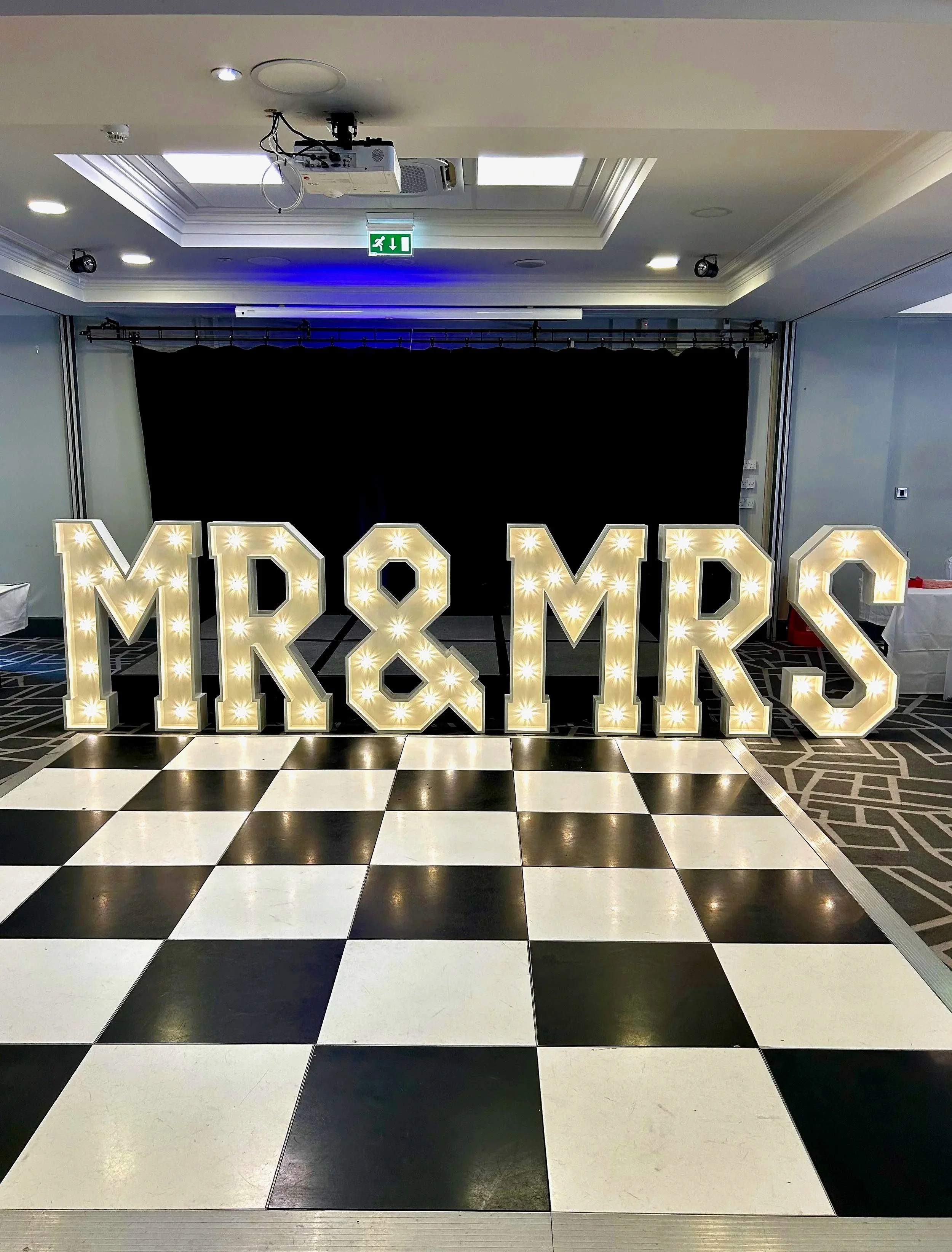 Large illuminated marquee letters spelling 'MR & MRS' on a checkered black and white flooring, with a black curtain backdrop and ceiling lights in the background.