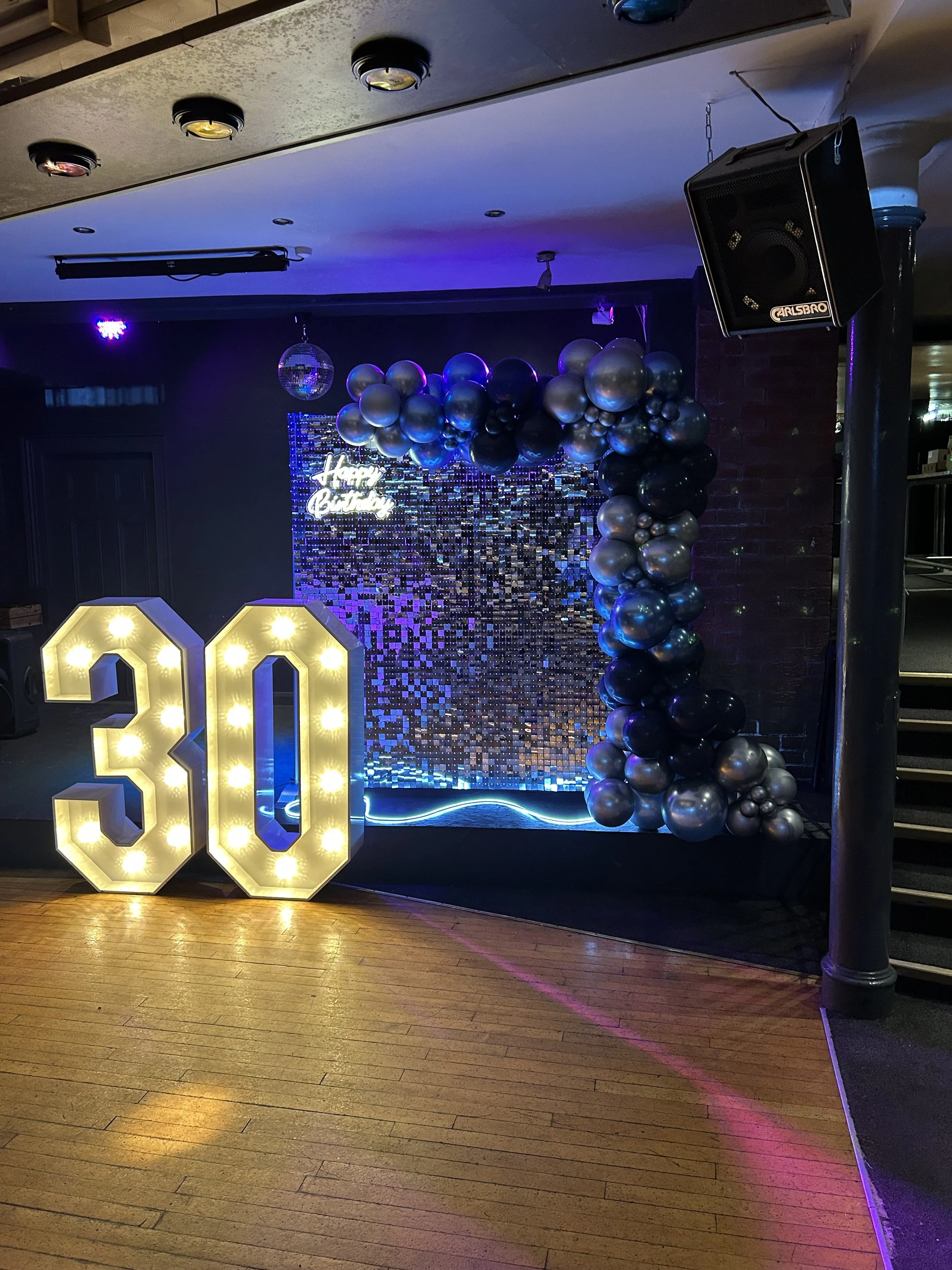 Decorative birthday backdrop with a large illuminated number 30, black, silver, and blue balloons, a small disco ball, and a 