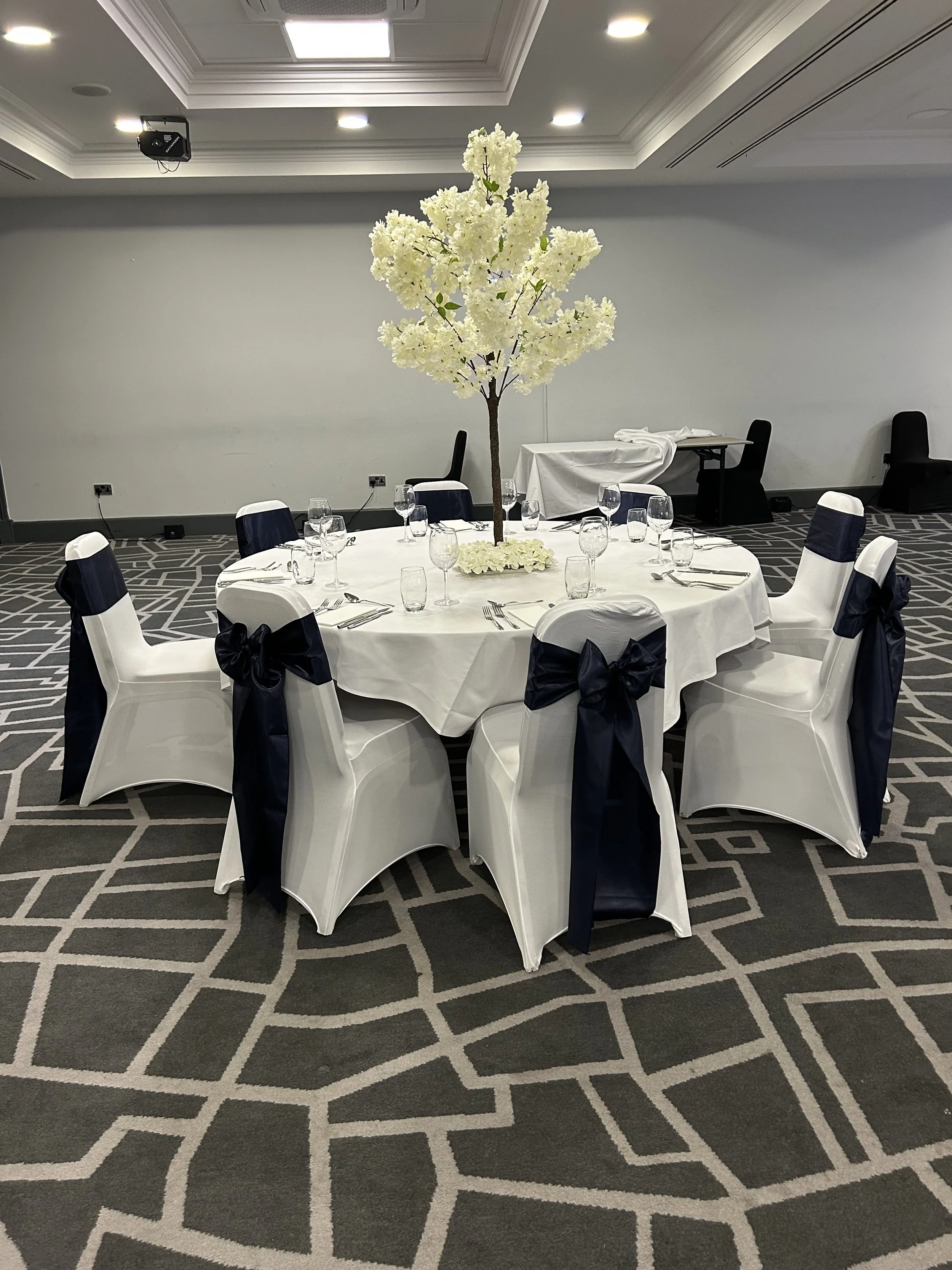 Round banquet table decorated with white tablecloth, surrounded by white chairs with navy blue sashes and bows, and centerpieces of white floral arrangements, in a banquet hall with patterned gray carpet and plain white walls.