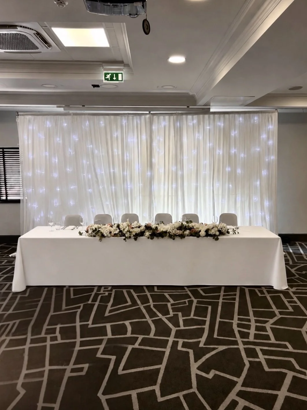 A wedding or event reception setup with a long white table, decorated with a floral centerpiece and wine glasses. Behind it is a backdrop of white curtains with soft lighting. The room has patterned carpeting and a drop ceiling with an exit sign and 