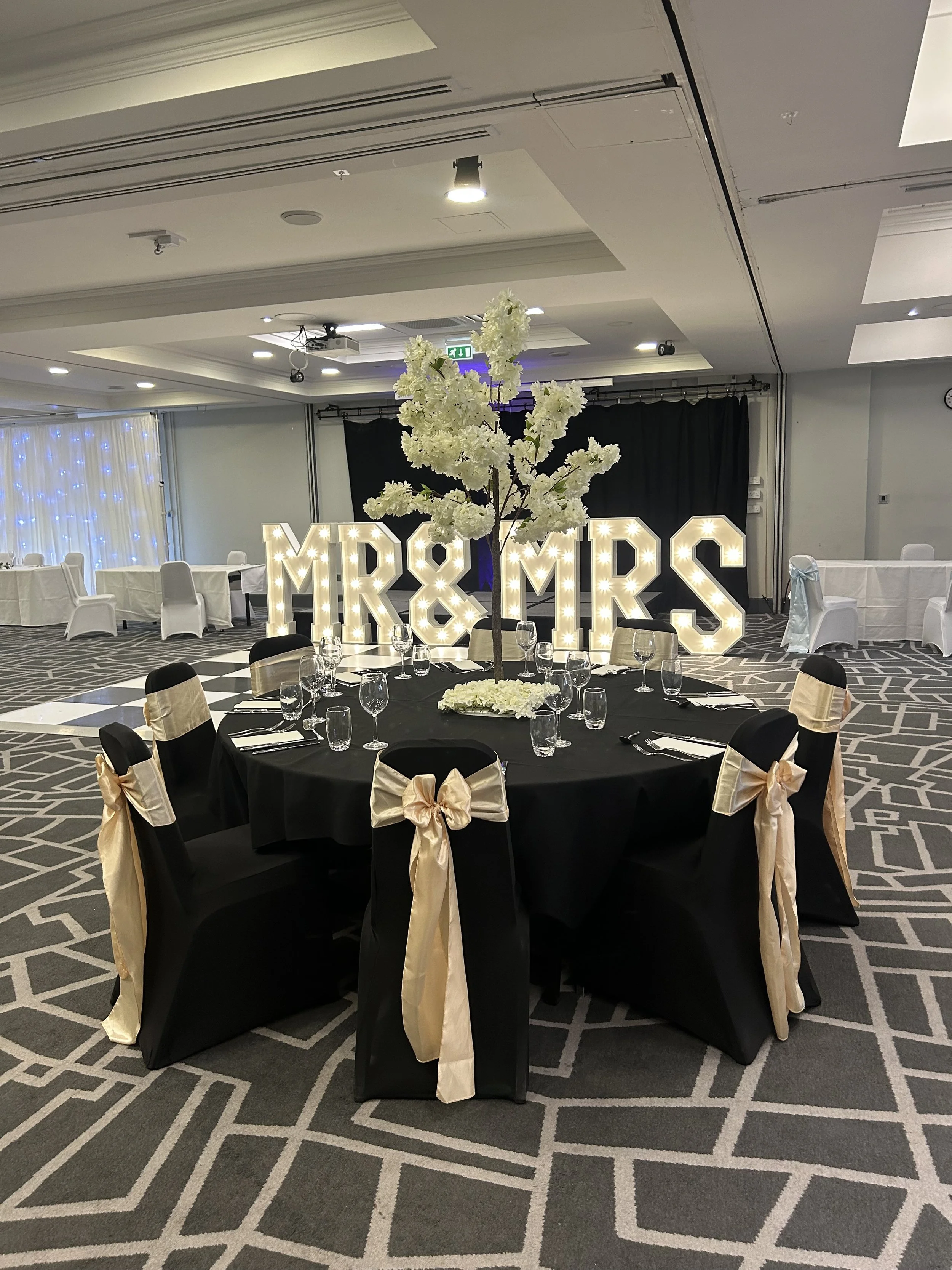 Wedding reception table with black tablecloth, gold and black chairs, champagne-colored sashes on chairs, centerpiece with white flowers and branches, large illuminated 