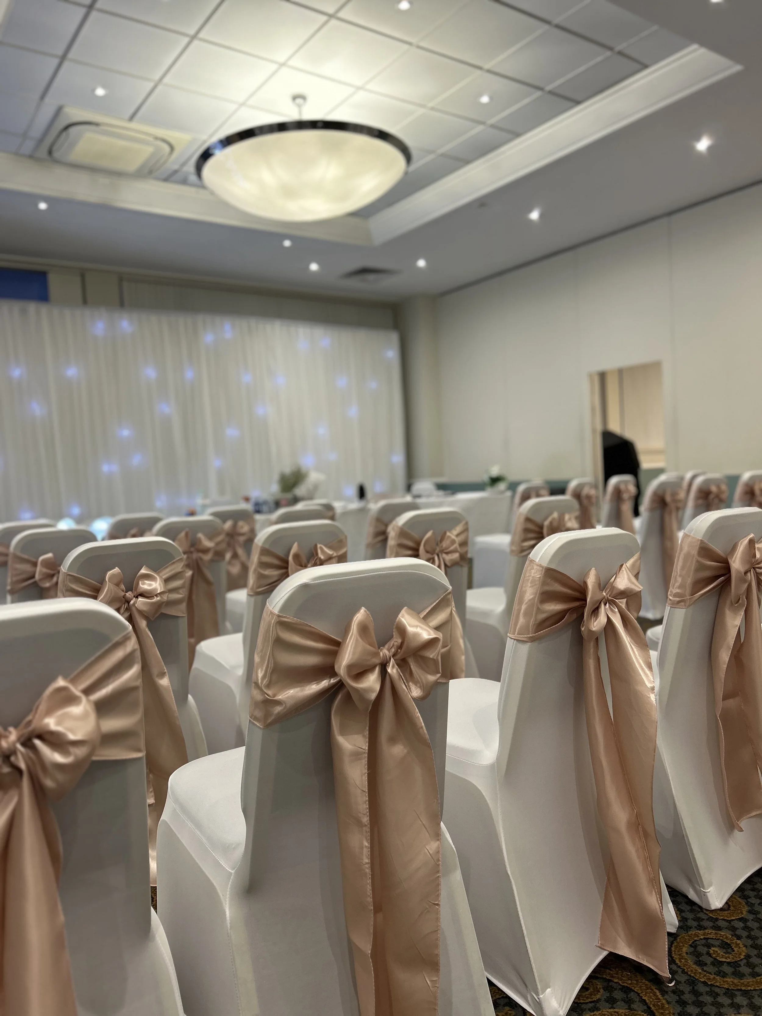 Empty banquet hall with white chairs decorated with beige satin bows, arranged in rows, with a stage area in the background featuring a curtain with small lights. The ceiling has a large round chandelier and recessed lighting.