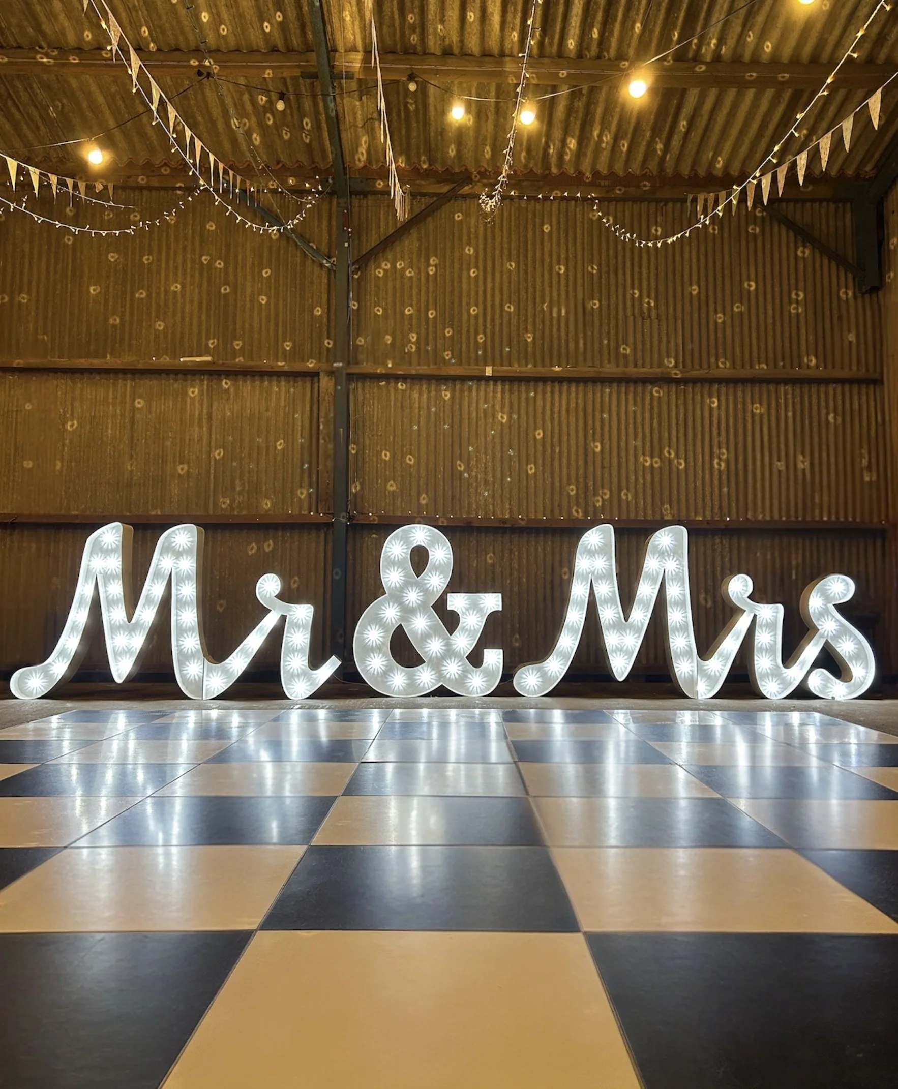 Decorative event space with string lights, bunting banners, a corrugated metal ceiling, and large illuminated letters on a checkered yellow and black tiled floor.