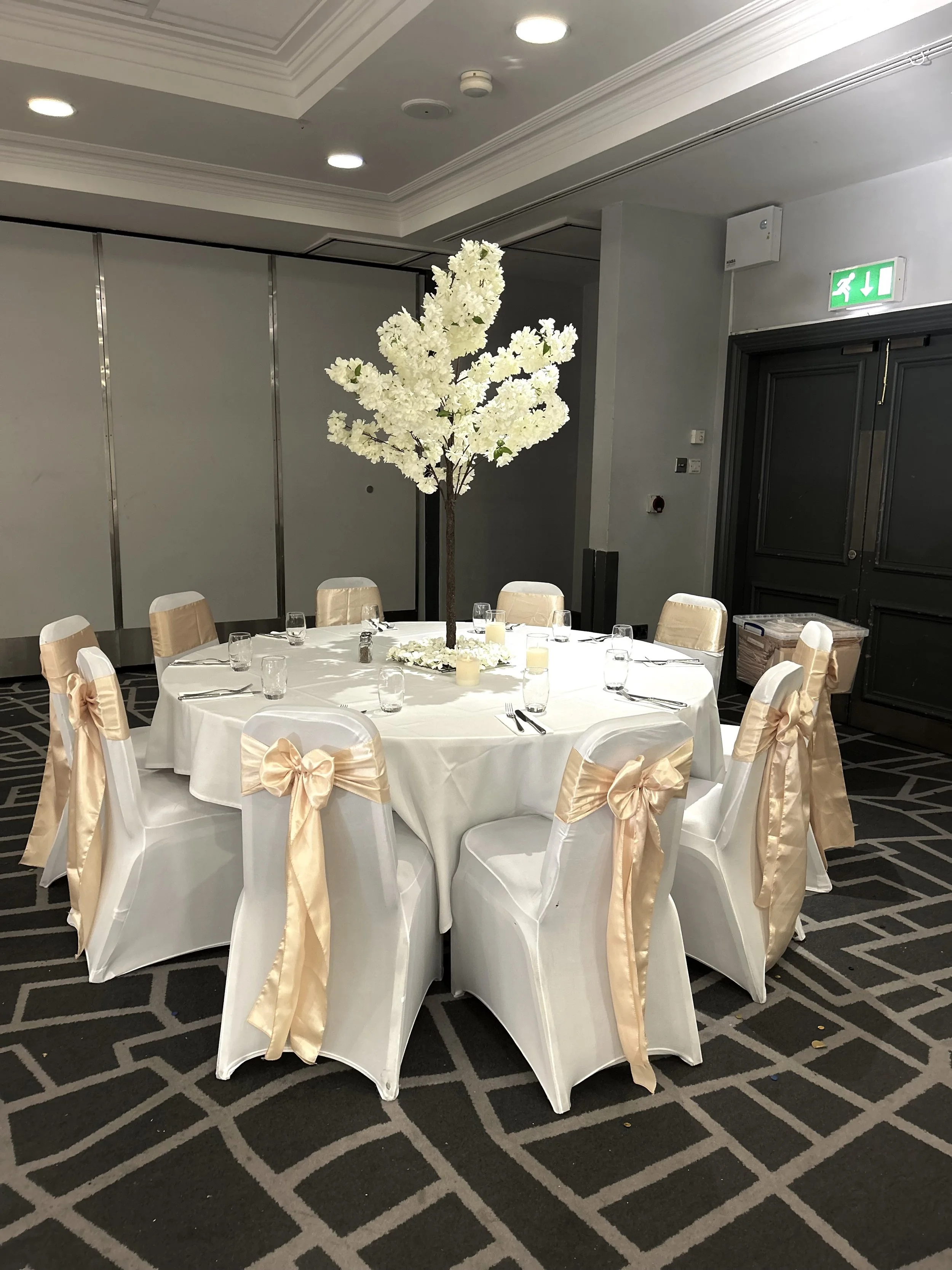 Round banquet table decorated with a tall white flower centerpiece and surrounded by chairs with cream-colored bows, set in a spacious, elegant room.