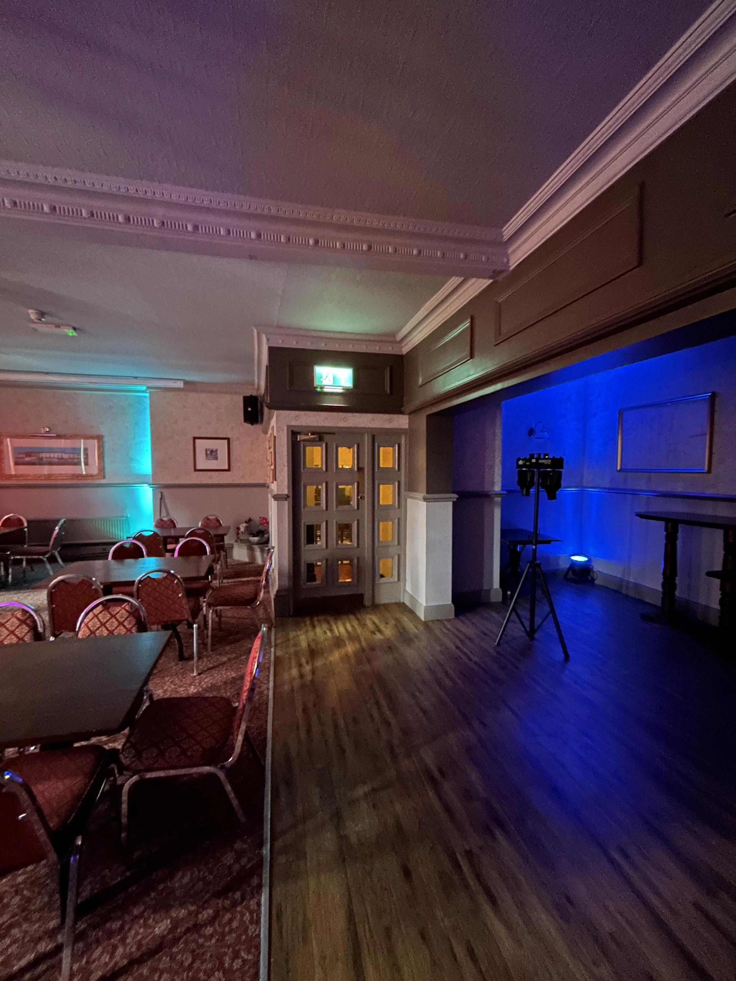 An empty event room with tables and chairs, some illuminated with colorful lights, and a small stage with lighting equipment.