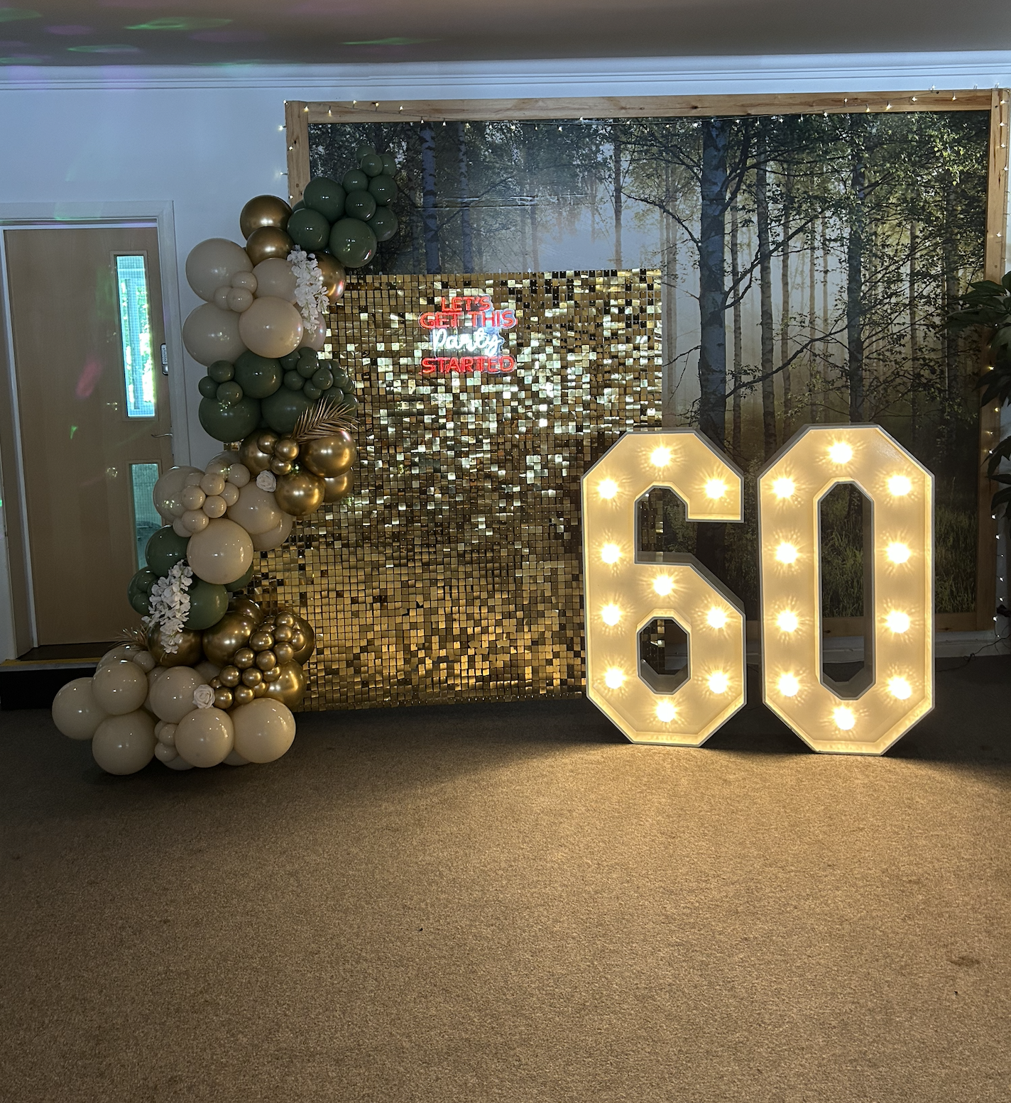 Decorative birthday celebration setup with large illuminated '60' sign, balloon arrangements in white, green, and gold, a shimmering gold backdrop, and a forest-themed mural in the background. A sign reads 'Let's get this party started'.
