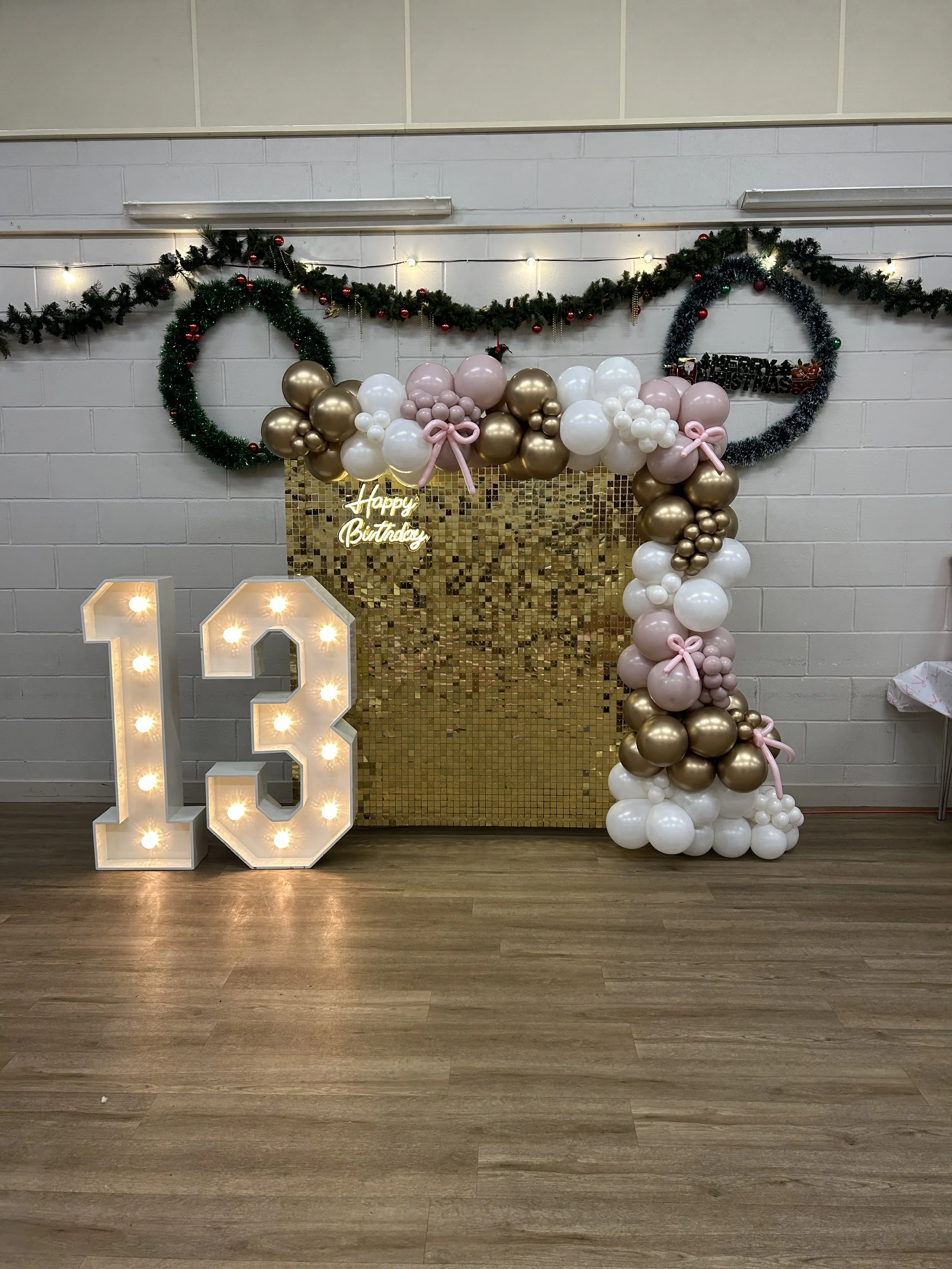 Decorative backdrop with gold, white, and pink balloons, a lit 'Happy Birthday' sign, and large illuminated '13' for a birthday celebration. Green garland with red ornaments and string lights above, situated on a brick wall, with a gold mosaic panel 