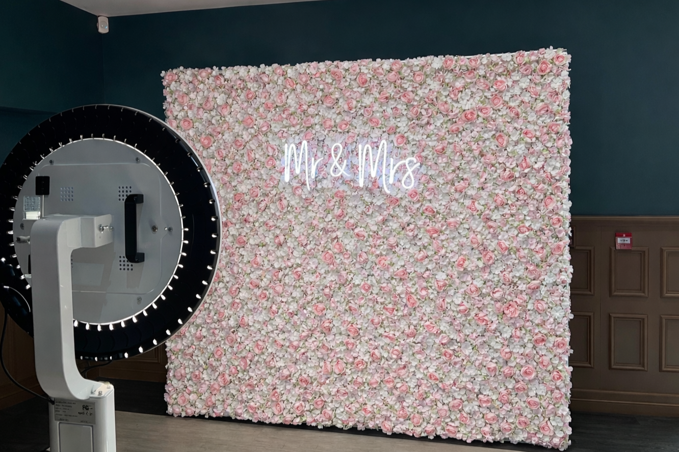 A floral backdrop with pink and white roses and greenery, with the words 'Mr & Mrs' in neon lights, and a photo booth prop machine in front.