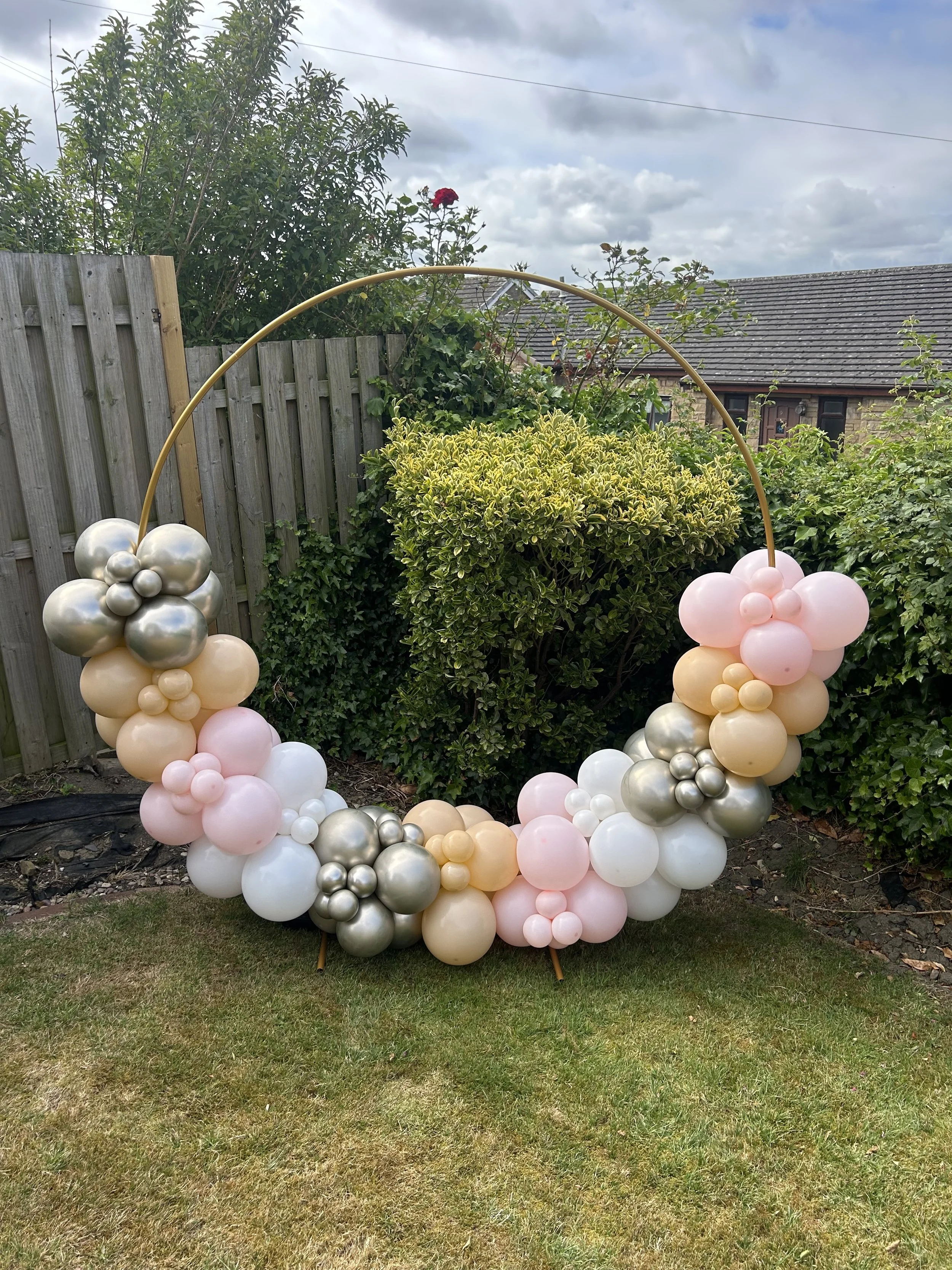 Decorative balloon arch with pastel pink, white, gold, and silver balloons arranged in clusters, set outdoors on a grass lawn with a wooden fence and green shrubbery in the background.