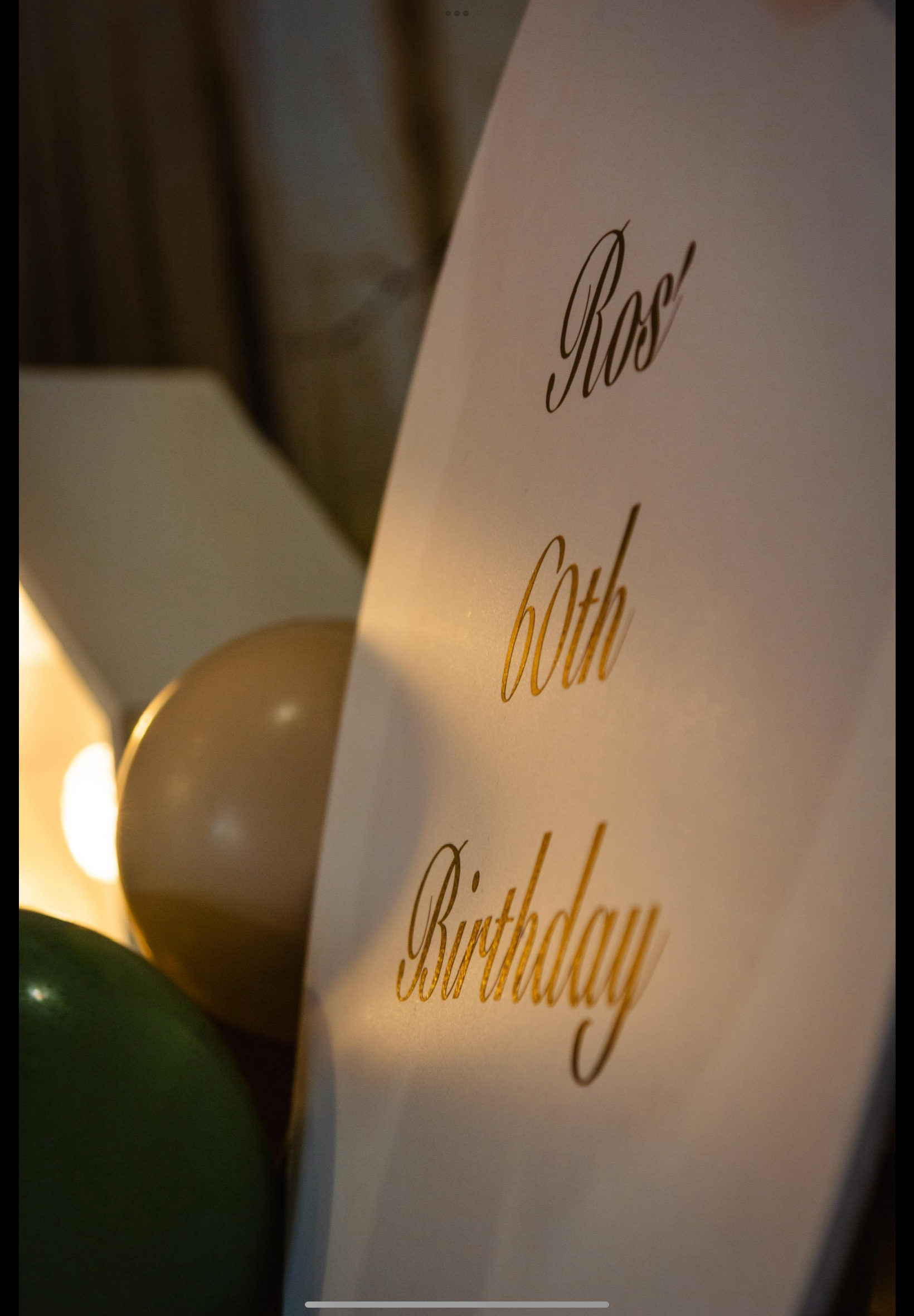 A white sign with gold lettering that reads "Rosa's 60th Birthday," surrounded by gold, white, and green balloons, and warm lighting.