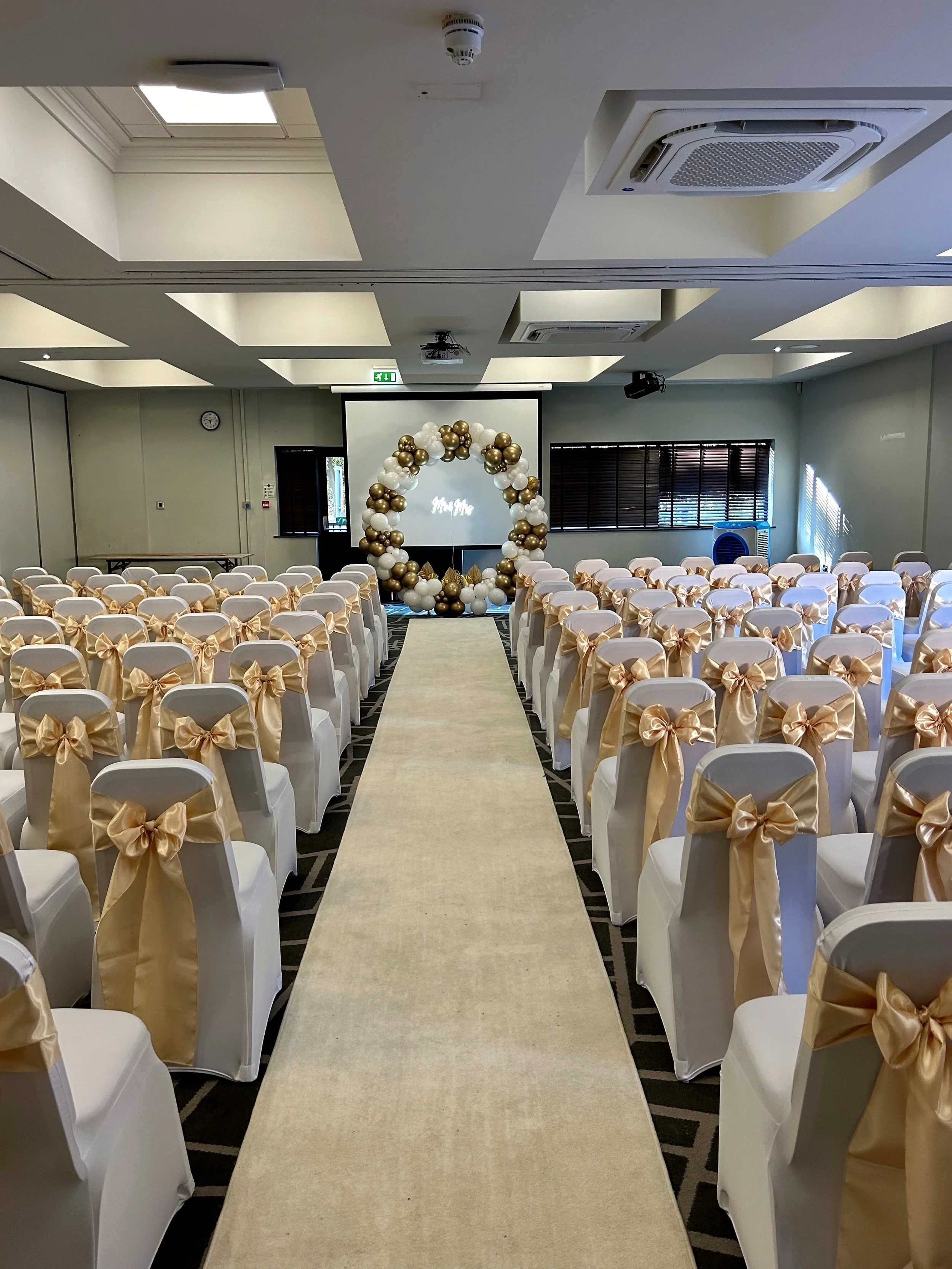 An indoor wedding reception area set with white chairs decorated with gold ribbons, arranged in rows along a central aisle. At the front, there is a large balloon arch made of white and gold balloons, with a projection screen behind it displaying som
