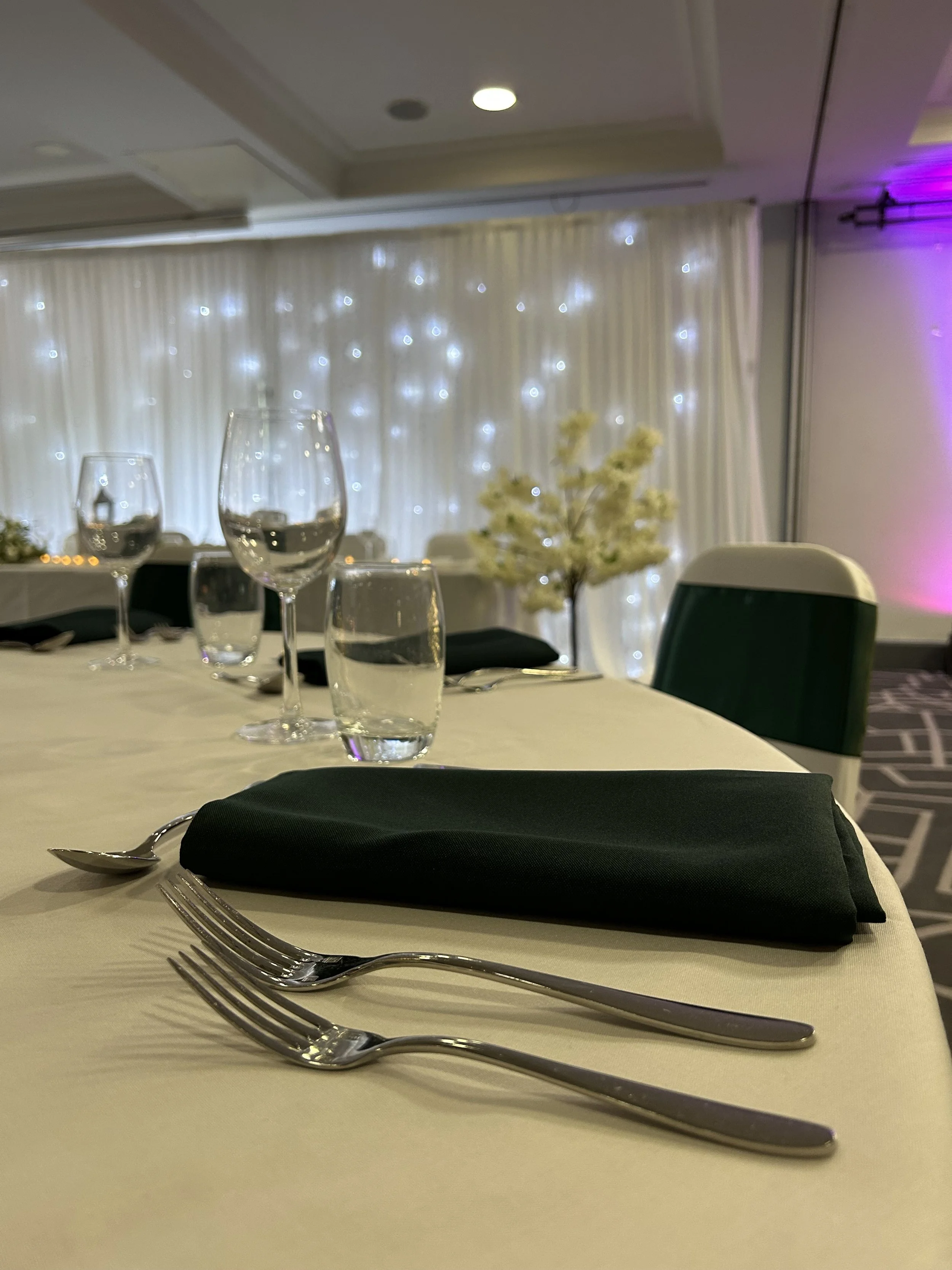 Elegant banquet table set with black cloth napkins, silver forks and spoons, wine glasses, water glasses, and a centerpiece with white flowers, in a decorated event hall with white curtains and string lights.