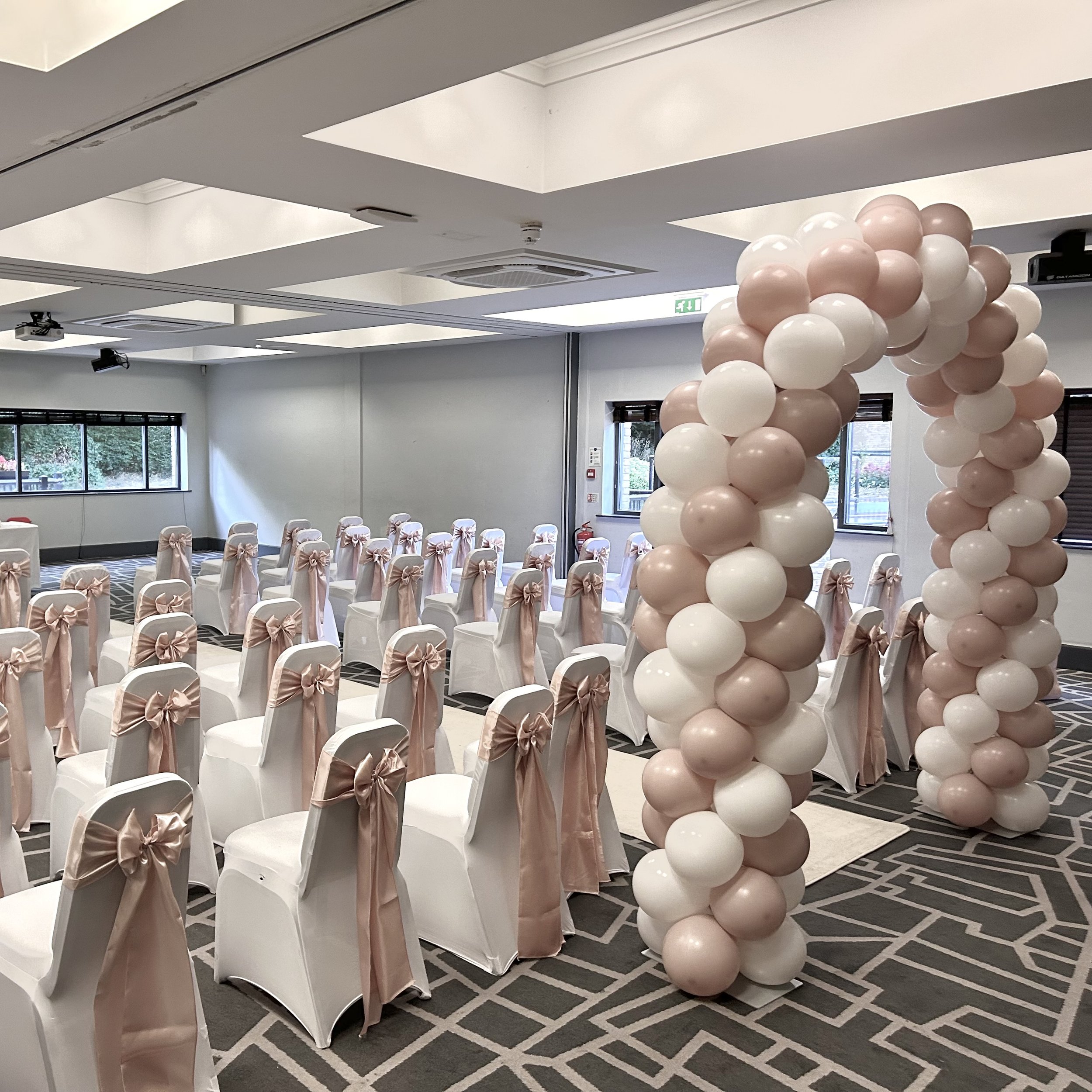 A decorated event space with 90 pink and white balloons forming columns, white chairs with pink ribbons and bows arranged in rows, and a large window in the background.