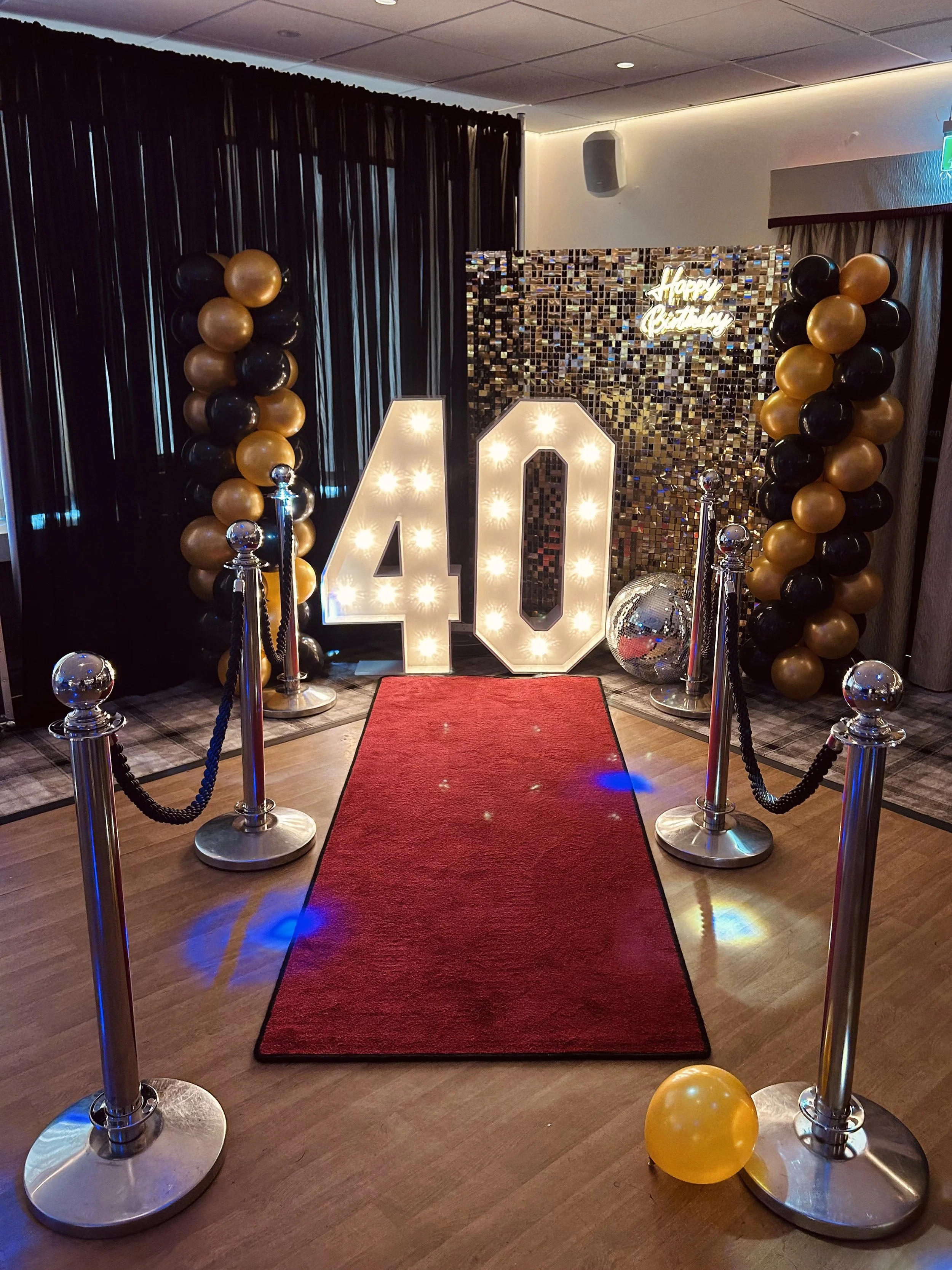 A birthday celebration setup with a red carpet, large illuminated number '40', balloons in black and gold, a disco ball, and a backdrop with a neon sign that says 'Happy Birthday'.