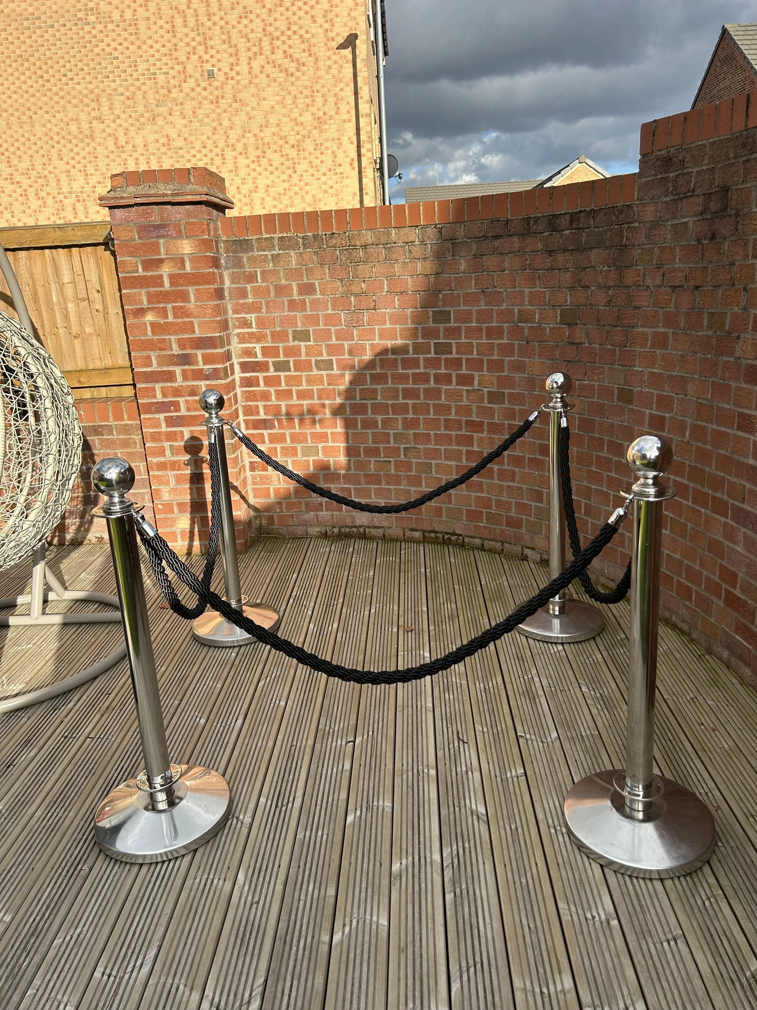 Outdoor wooden deck with metal stanchions and black ropes creating a barrier, brick wall with surrounding buildings in the background, cloudy sky overhead.
