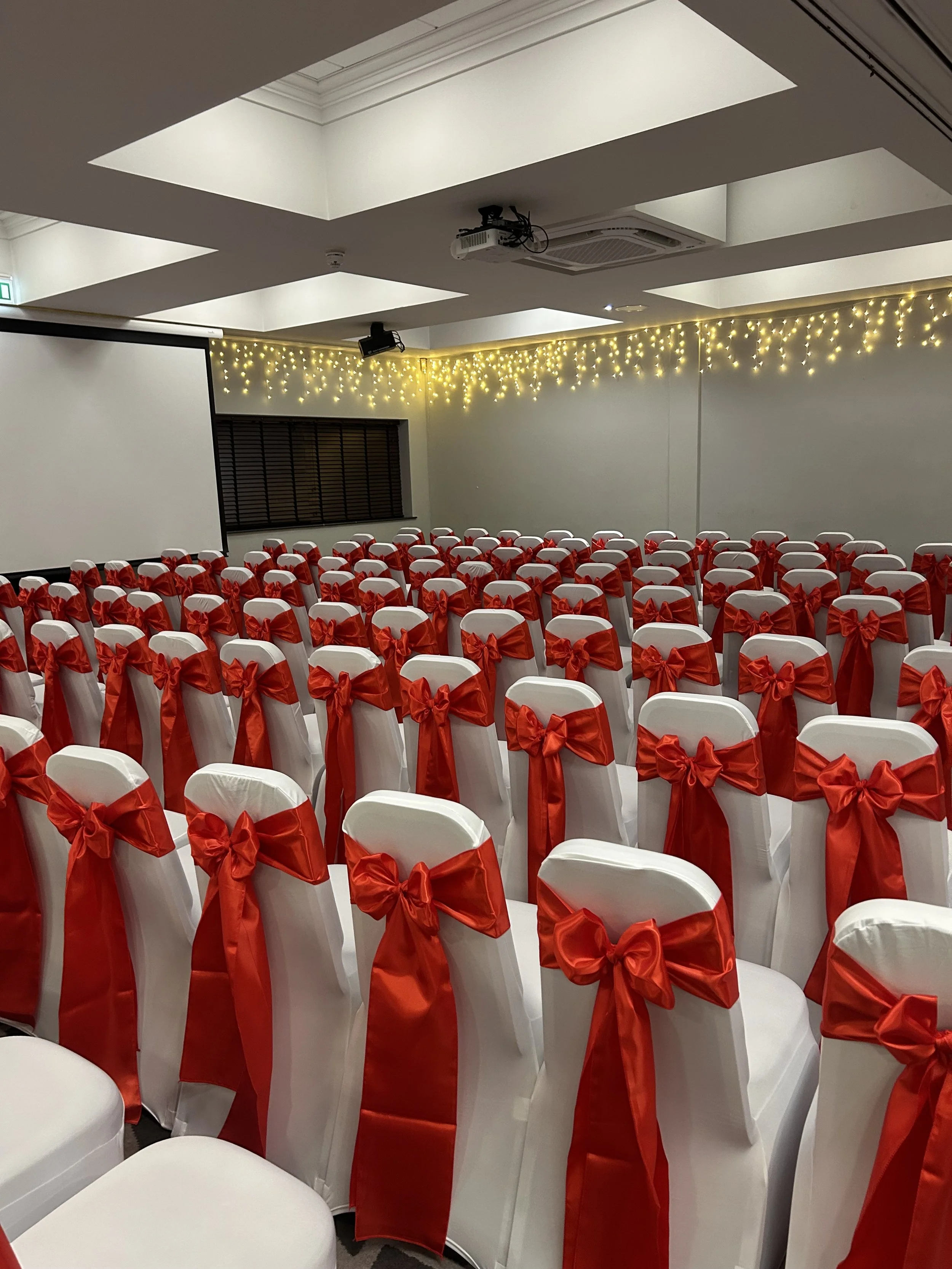 Rows of white chairs with red bows in a decorated room, likely set up for a wedding or event, with fairy lights hanging along the ceiling.