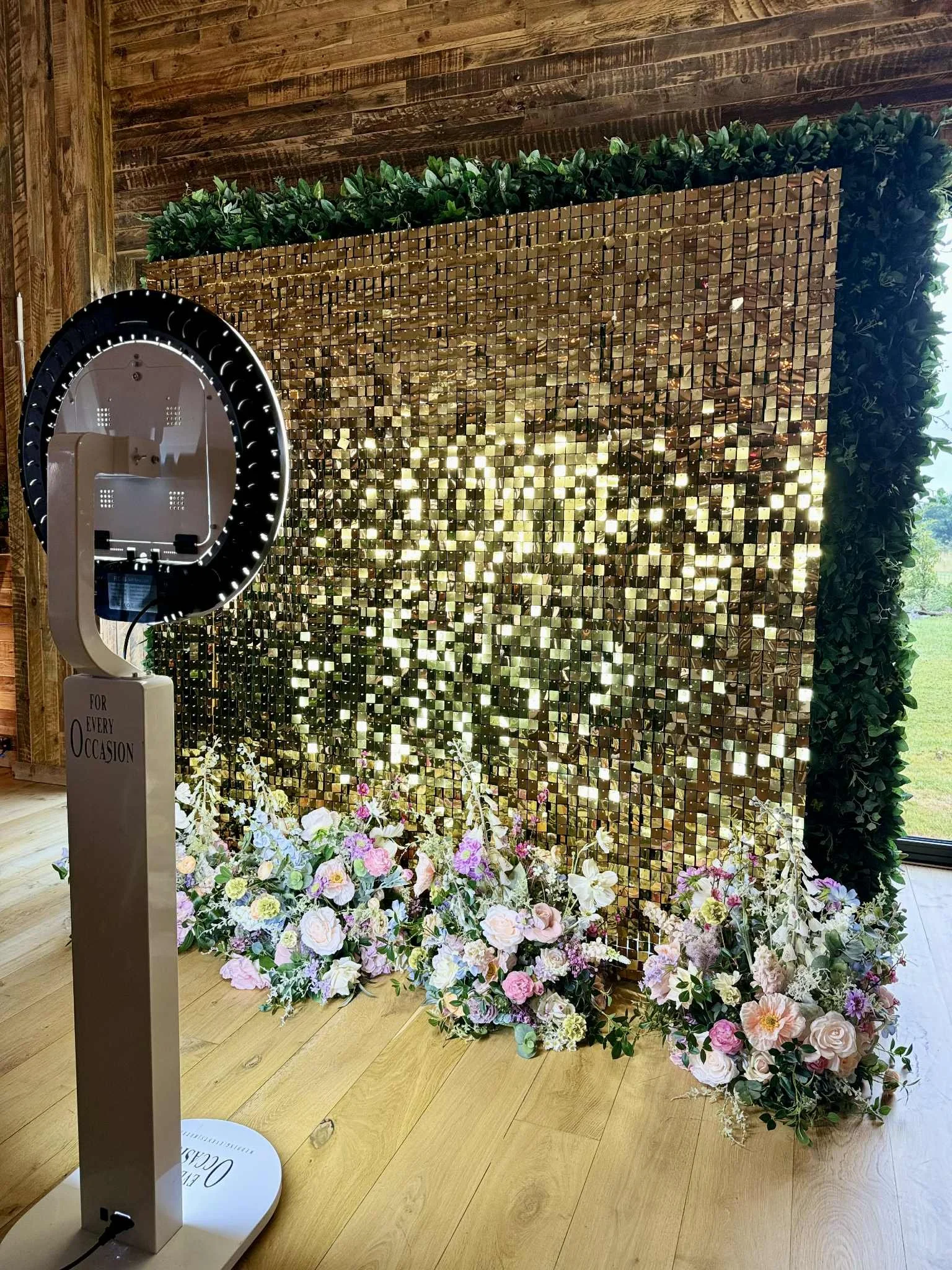 A photo booth backdrop consisting of a golden sequined wall, surrounded by green foliage at the top and sides, with floral arrangements of pink, white, and purple flowers at the base. There is a stand with a sign that reads 'For Every Occasion' and a