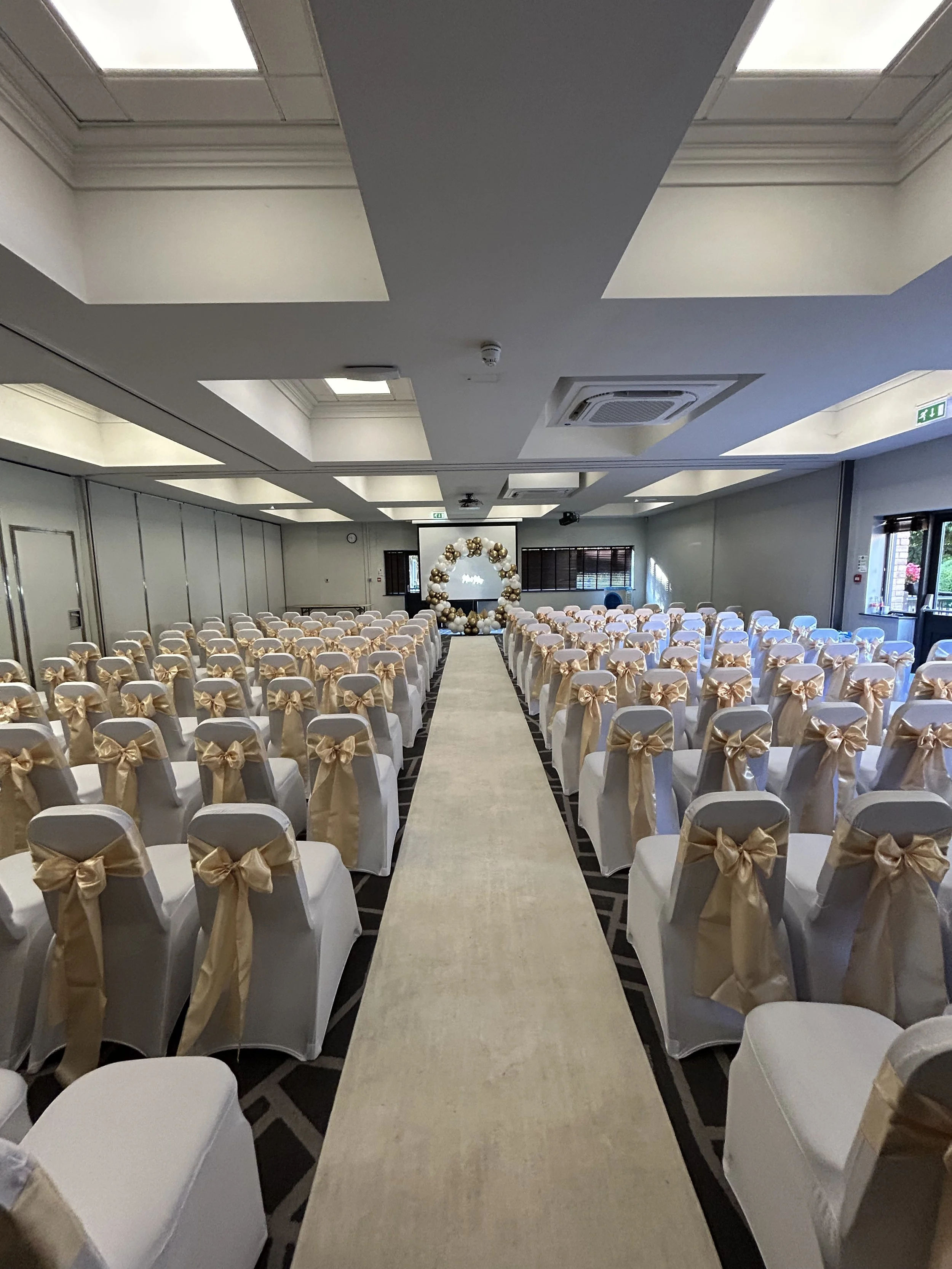 An elegant wedding ceremony setup with rows of white chairs decorated with gold sashes tied into bows, arranged on either side of an aisle leading to a floral arch at the front of the room.