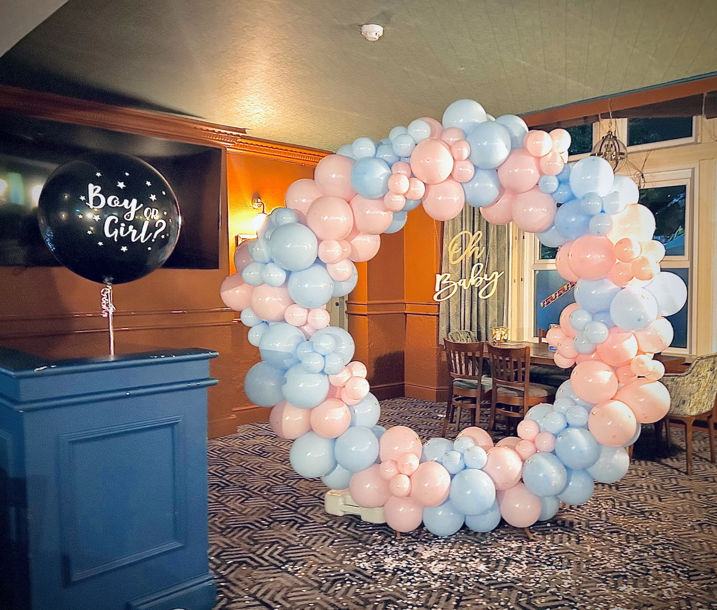 Decorative balloon arch in pastel pink and blue colors with a sign saying "Oh Baby" and a black balloon on a blue stand with "Boy or Girl?" written on it, set up for a gender reveal party in a room with wooden furniture and windows.