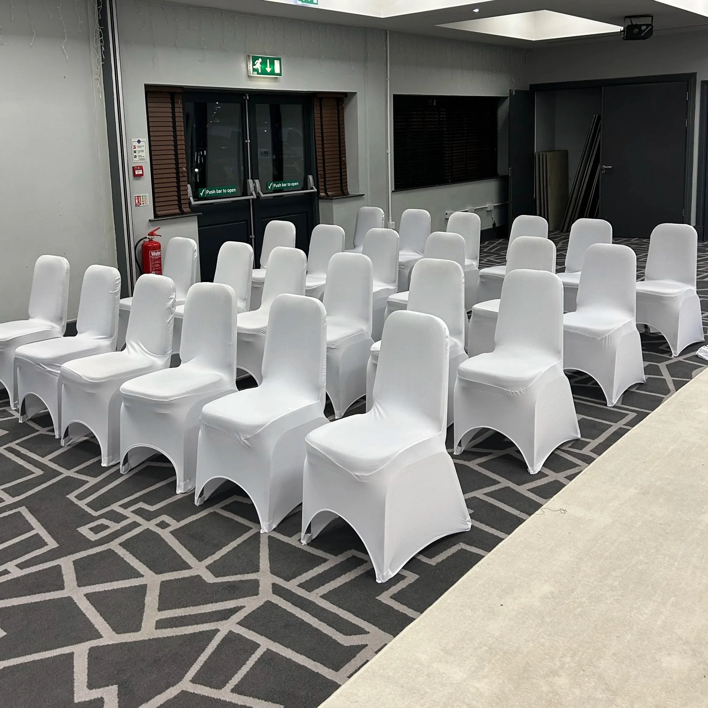 An indoor room with white covered chairs arranged in rows, black and white patterned carpet, and a door with an emergency exit sign above it.