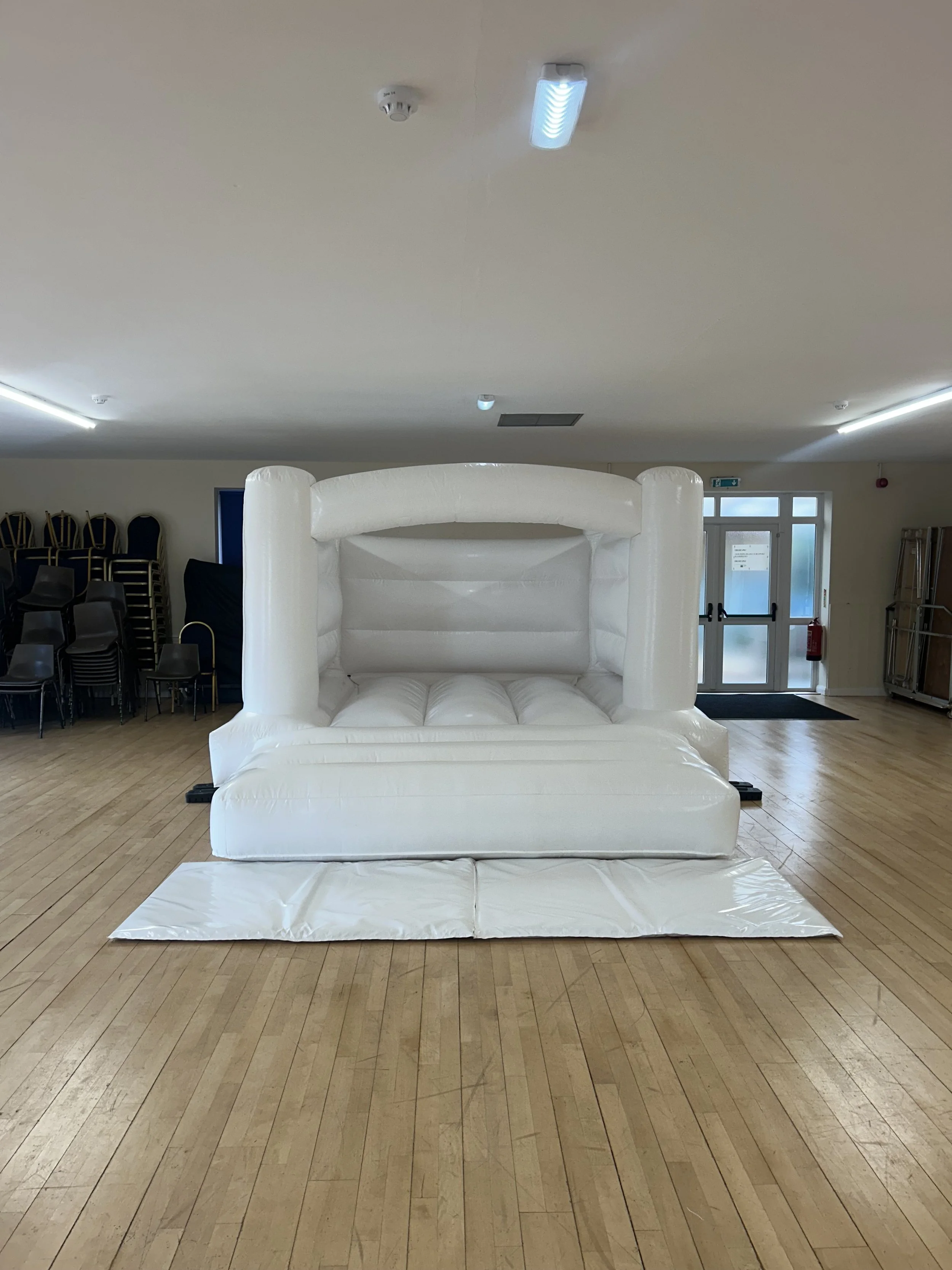 Indoor space with an inflatable white structure resembling a small castle or play area on a wooden floor. Chairs are stacked in the background on the left side, and there's a door with windows and a fire extinguisher on the right side.