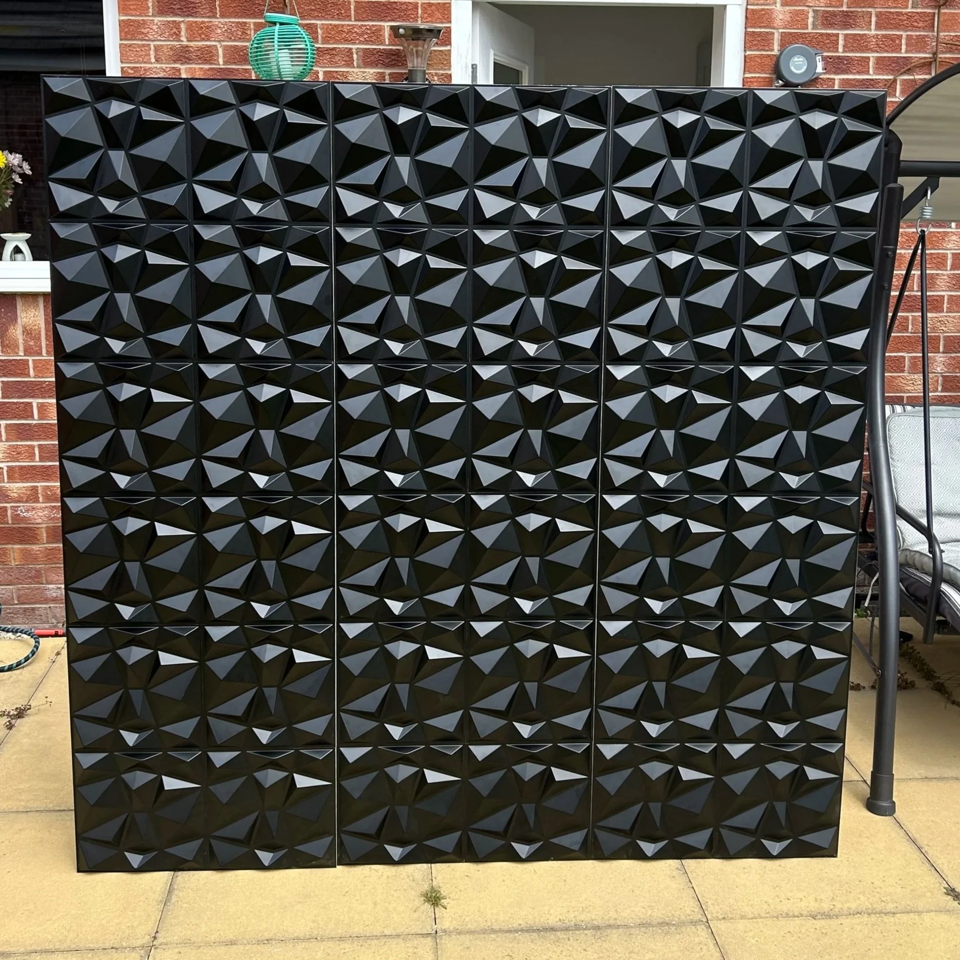 A large black decorative room divider with geometric patterns, standing outdoors on a patio with brick house wall, plants, and furniture in the background.