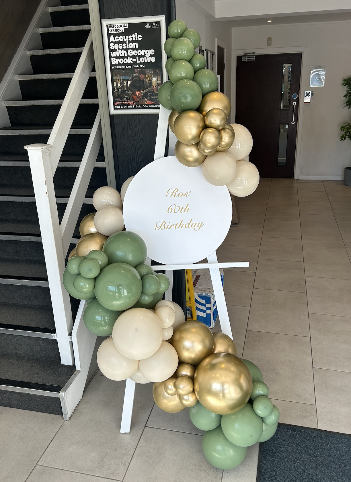 Decorative balloon arrangement with green, cream, and gold balloons surrounding a white sign that reads "Ros' 60th Birthday," placed on an easel near a staircase inside a building.